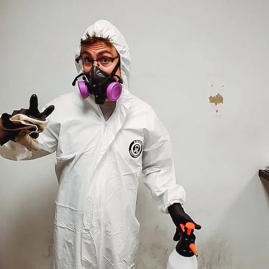 Trauma cleaner wearing protective hazmat suit, goggles, and a mask with purple filters, holding a spray bottle, making a peace sign with one hand.