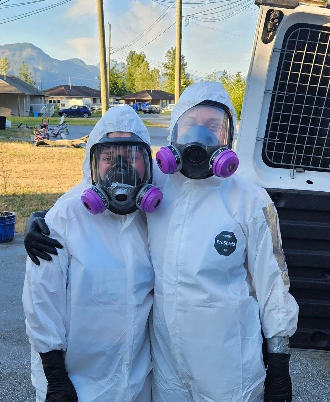 Two trauma cleaners dressed in full white protective suits with face shields and purple respirator masks, standing outdoors with a bright sky and mountains in the background.