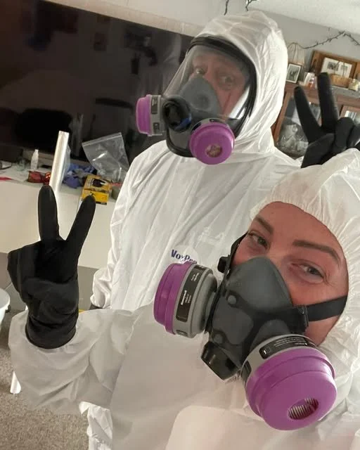 Two biohazard cleaners wearing full protective suits and masks with double stack cartridges, making peace signs.