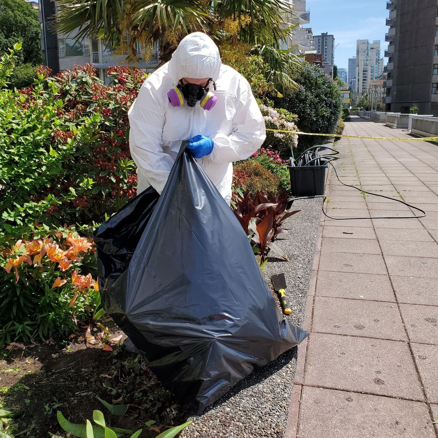 A biohazard cleaner in protective white suit, gloves, and a respirator mask is picking up trash with a black trash bag. They are working in an outdoor urban garden area with plants and flowers, surrounded by tall buildings.