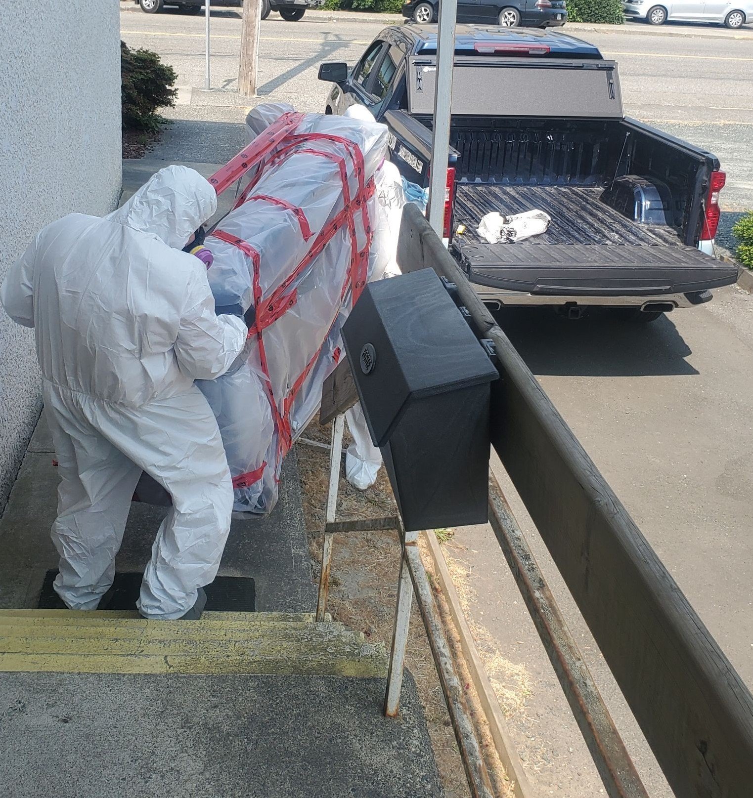 Two trauma cleaners dressed in white protective suits handling a sealed mattress wrapped in plastic and red tape outdoors, next to a railing and black trash bin, with pickup truck parked nearby.