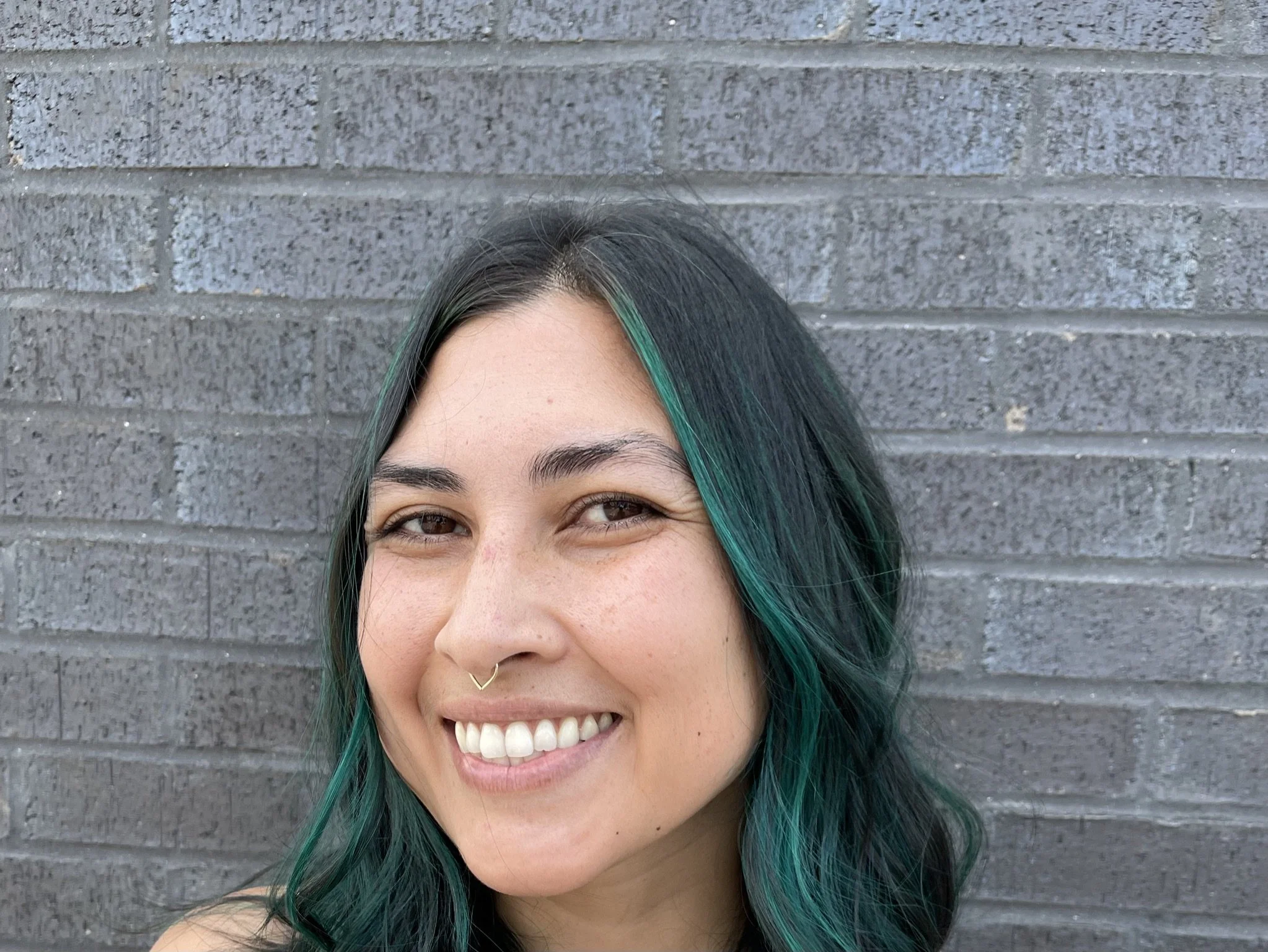 A young woman with black hair and green highlights smiling in front of a gray brick wall.