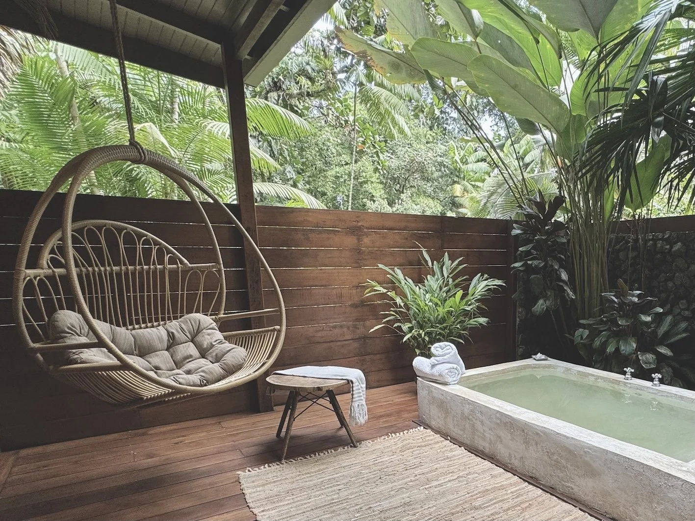 Junior Suite El agua: Outdoor tropical patio with hanging wicker chair, small wooden table with towel, concrete hot tub, lush green plants, and wooden privacy fence, overlooking dense forest.