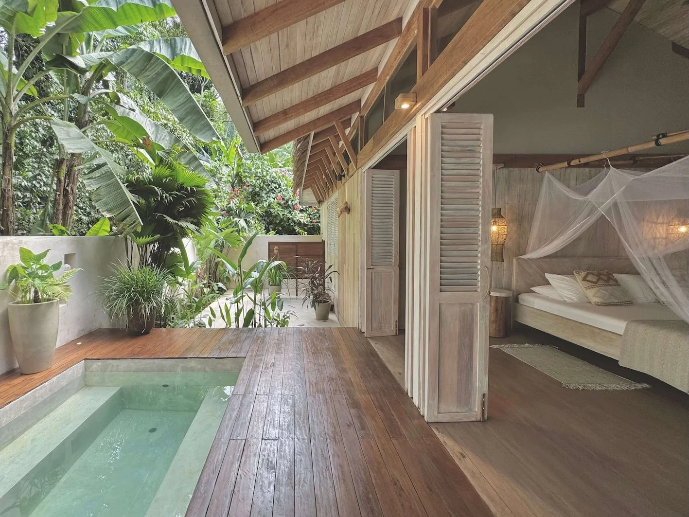 Master Suite La Selva: Open bedroom with mosquito net, wooden walls and flooring, connected to an outdoor patio with small pool, lush tropical plants, and wooden deck.