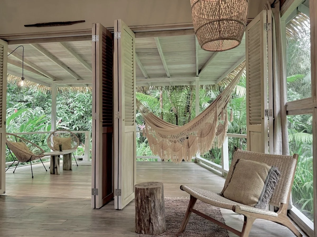 Garden Suites: El Cielo or La Tierra: Covered outdoor patio with wicker chairs, a hammock, a wooden side table, and lush green tropical plants outside.