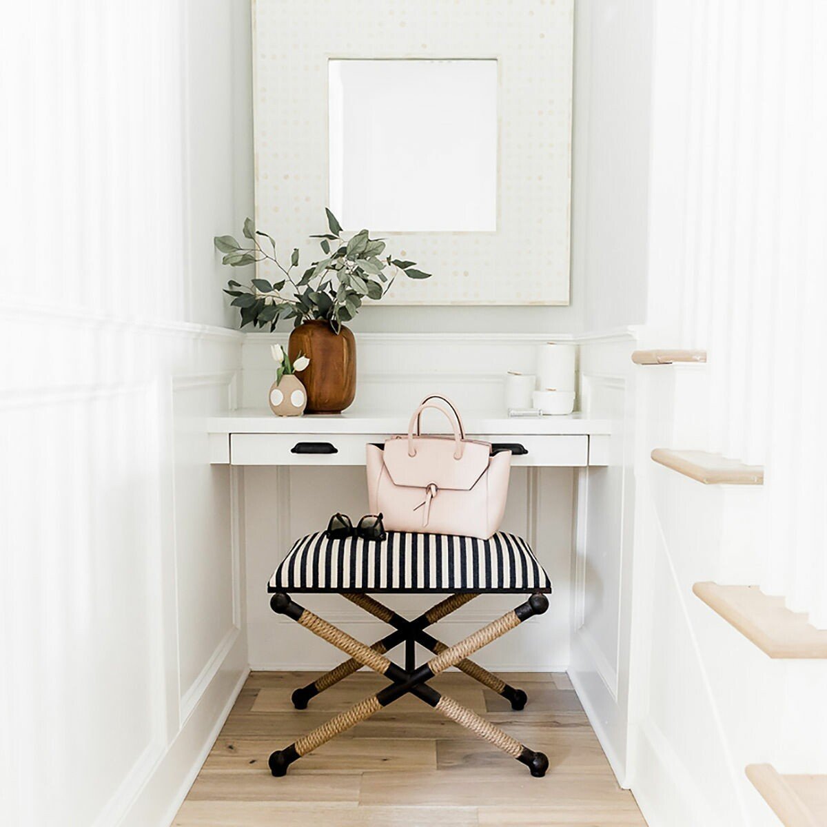 A small entryway with a white wooden wall, staircase, and a dark wooden and striped fabric bench. Decorative items on the white desk include a vase with green leaves, a small vase with white flowers, a pink handbag on the bench, and a pair of sunglas