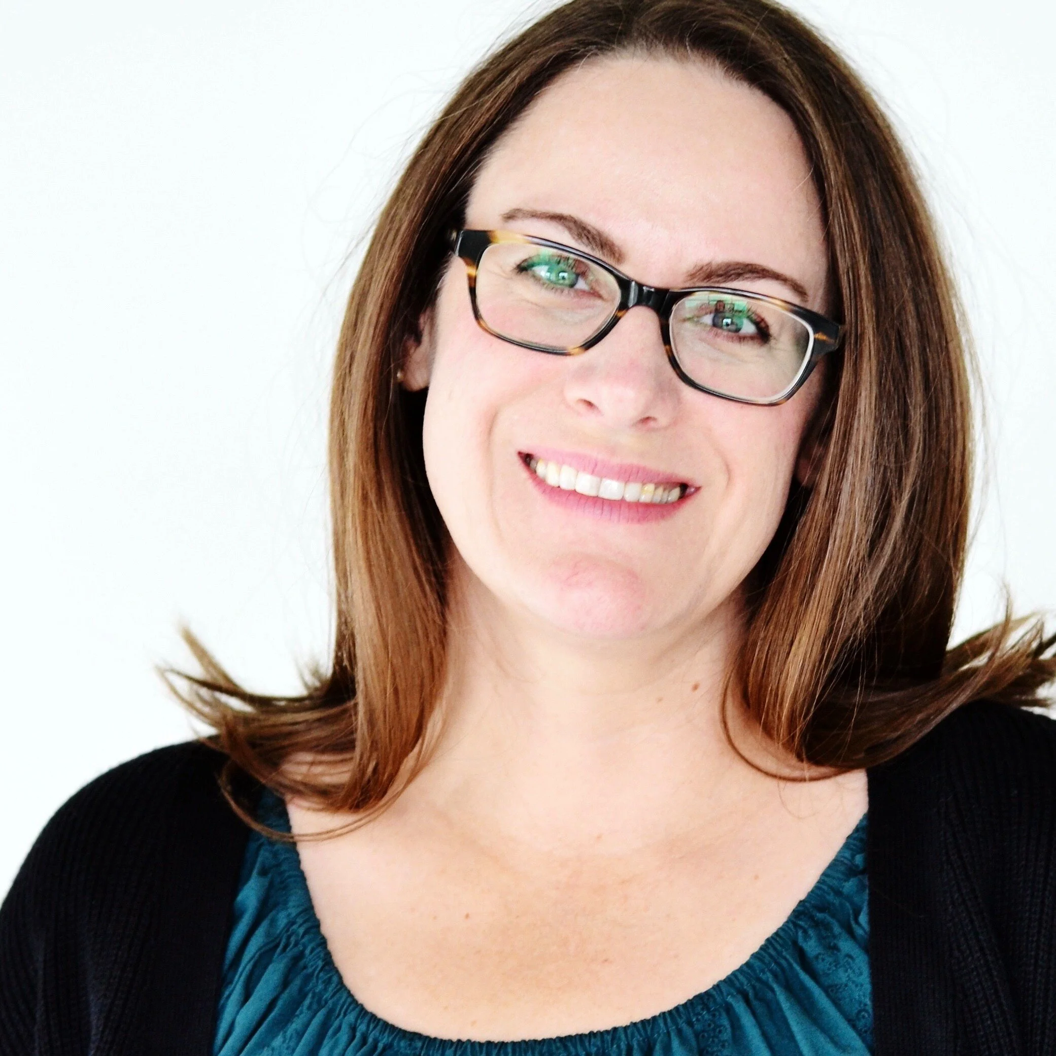 A woman with shoulder-length brown hair, glasses, and a smile, wearing a teal top and black cardigan, standing against a white background.