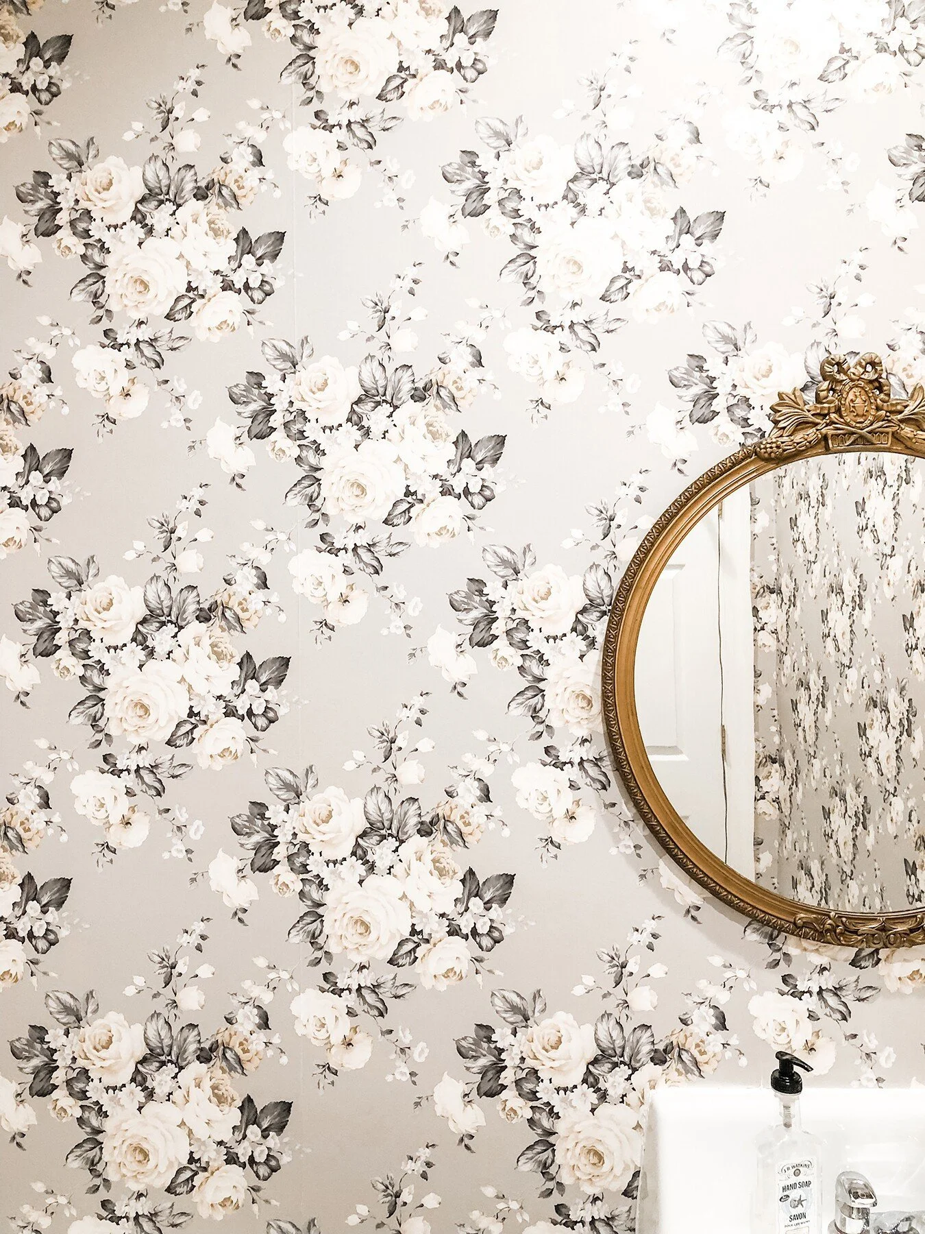 A bathroom wall with a floral wallpaper featuring white and gray roses and leaves, a gold-framed oval mirror, and a white sink with a hand soap dispenser.