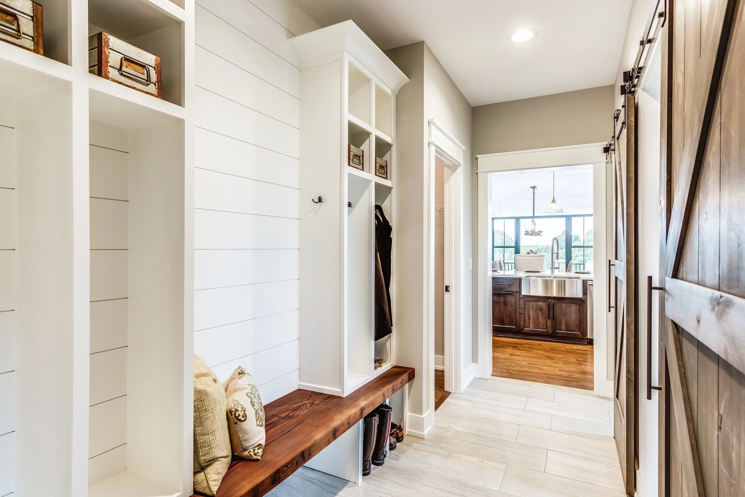 A modern entryway with a built-in wooden bench, white shelving, and hooks, leading into a kitchen with wooden cabinets and a large window.