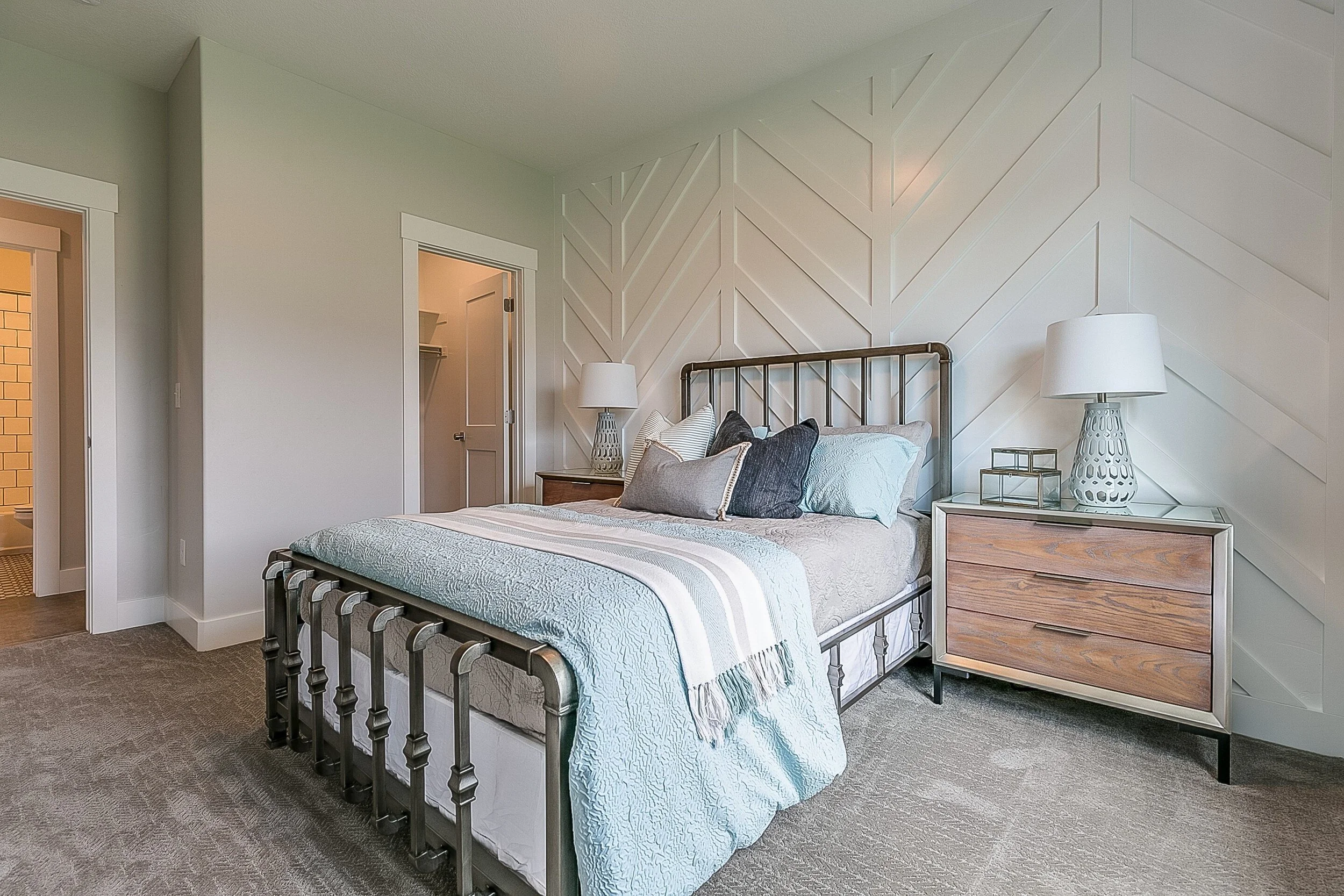 A modern bedroom with a metal bed frame, light bedding, and decorative pillows. Two bedside tables with lamps, and a textured accent wall behind the bed. An open door reveals a walk-in closet.