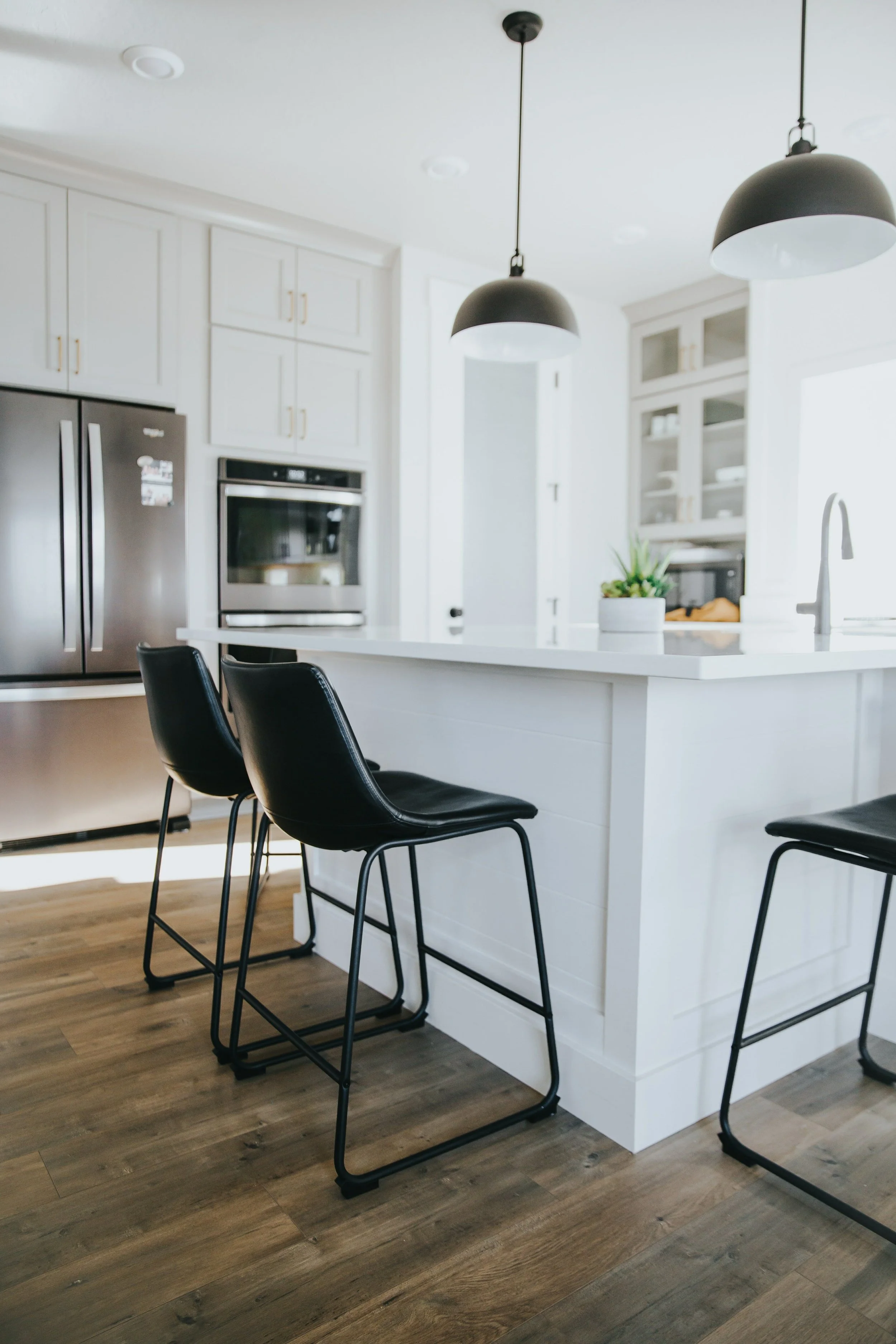 Modern kitchen with white cabinets, stainless steel appliances, black bar stools, pendant lights, and wooden flooring.