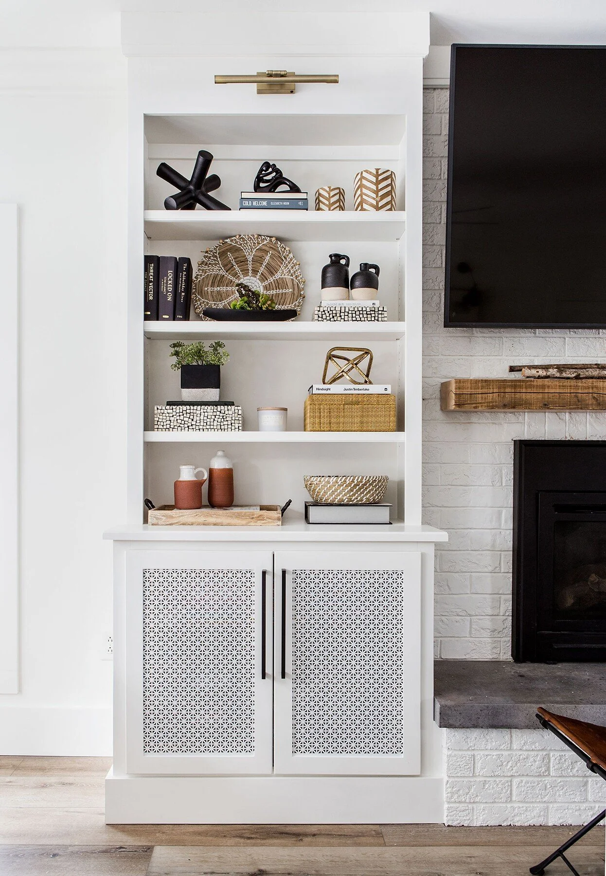 White built-in bookshelf with decorative items and books, adjacent to a brick fireplace with a wooden mantel and a large television mounted on the wall.