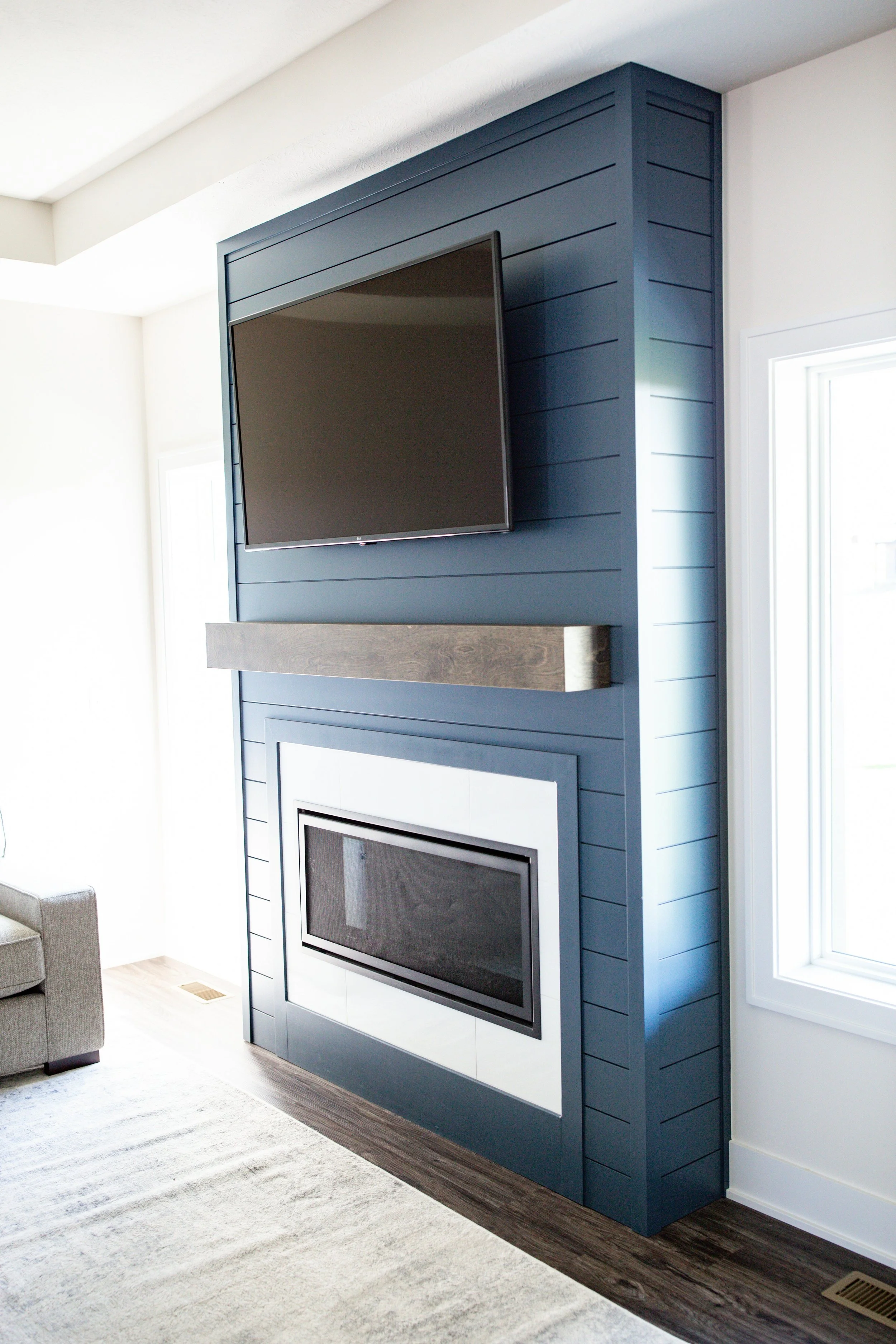Interior view of a living room with a blue shiplap accent wall featuring a mounted flat-screen TV, a modern fireplace, and a wooden mantel. A window to the right provides natural light, with a part of a sofa visible on the left and a beige area rug on the floor.