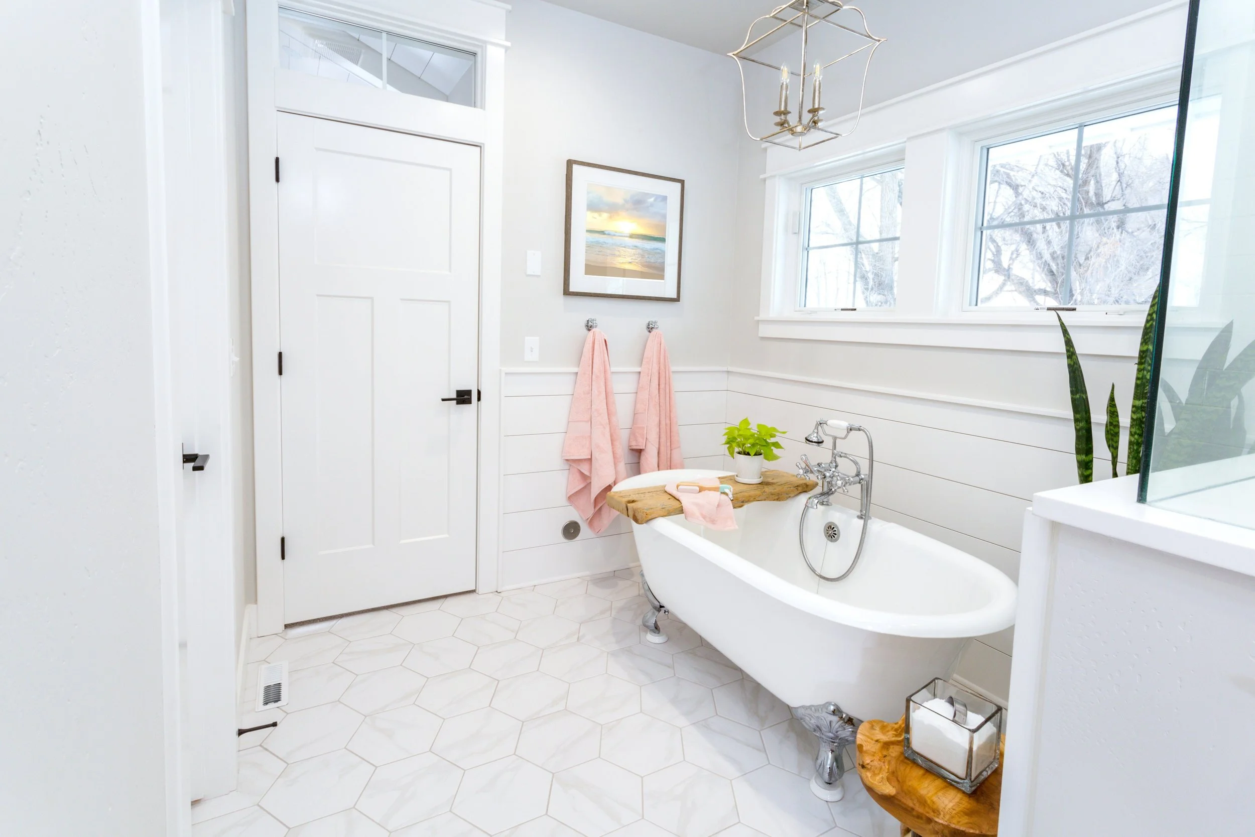 A bright bathroom with white hexagonal tile flooring, a clawfoot bathtub with a wood tray, pink towels, a plant, and a window with snowy outdoor scenery.