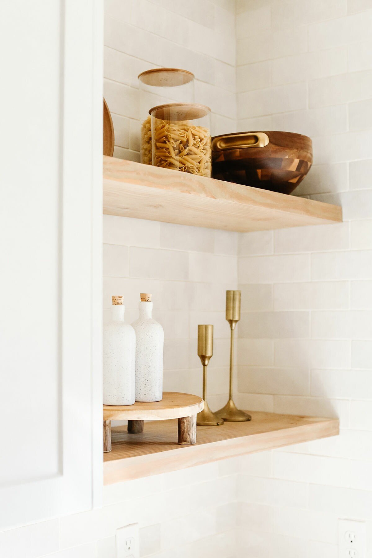 Wooden shelves with glass jars containing pasta, a wooden bowl, white bottles, and brass candle holders on a tiled wall.