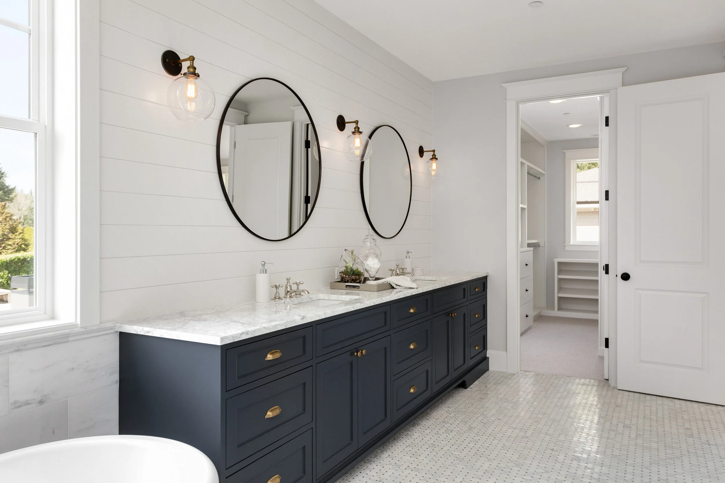 Bathroom with double vanity, navy blue cabinets with gold hardware, marble countertop, two oval mirrors, two wall-mounted light fixtures, white shiplap walls, and walk-in closet visible through an open door.