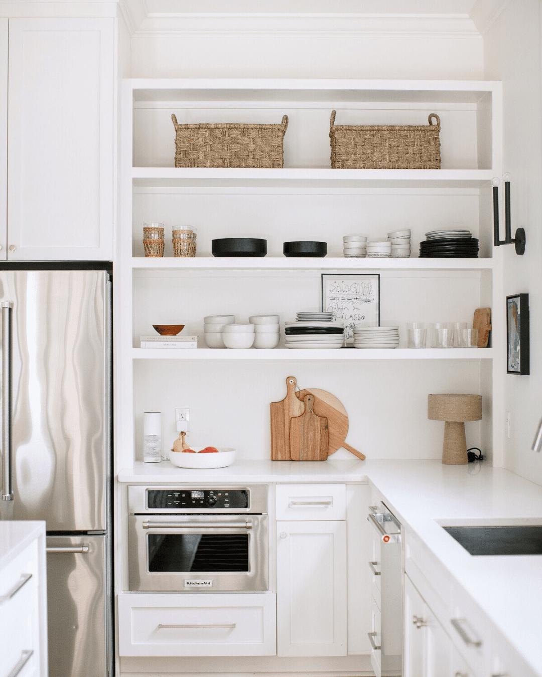 Open kitchen shelf with woven baskets, glassware, black and white dishes, and framed art, adjacent to a refrigerator and oven.
