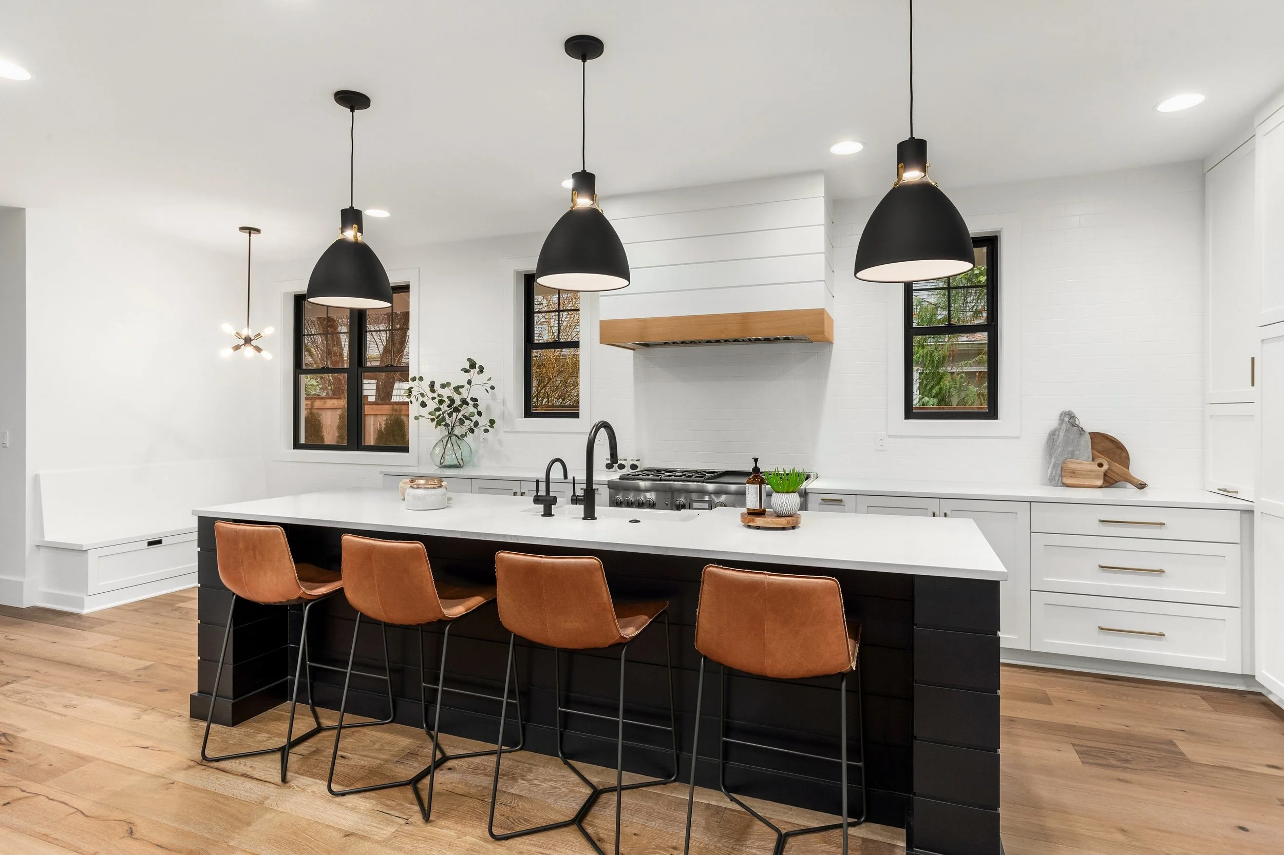 Modern kitchen with black pendant lights over a white island with five brown bar stools, white cabinetry, black window frames, and wooden accents.