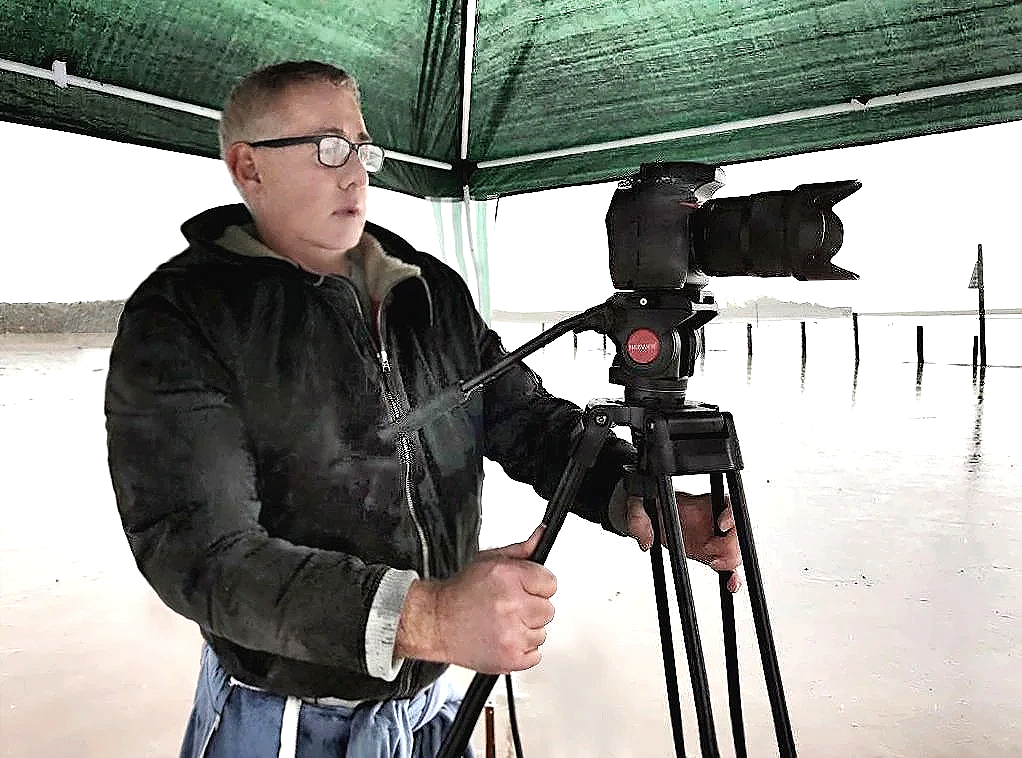 Young man with glasses operating a camera on a tripod beneath a green canopy outdoors.