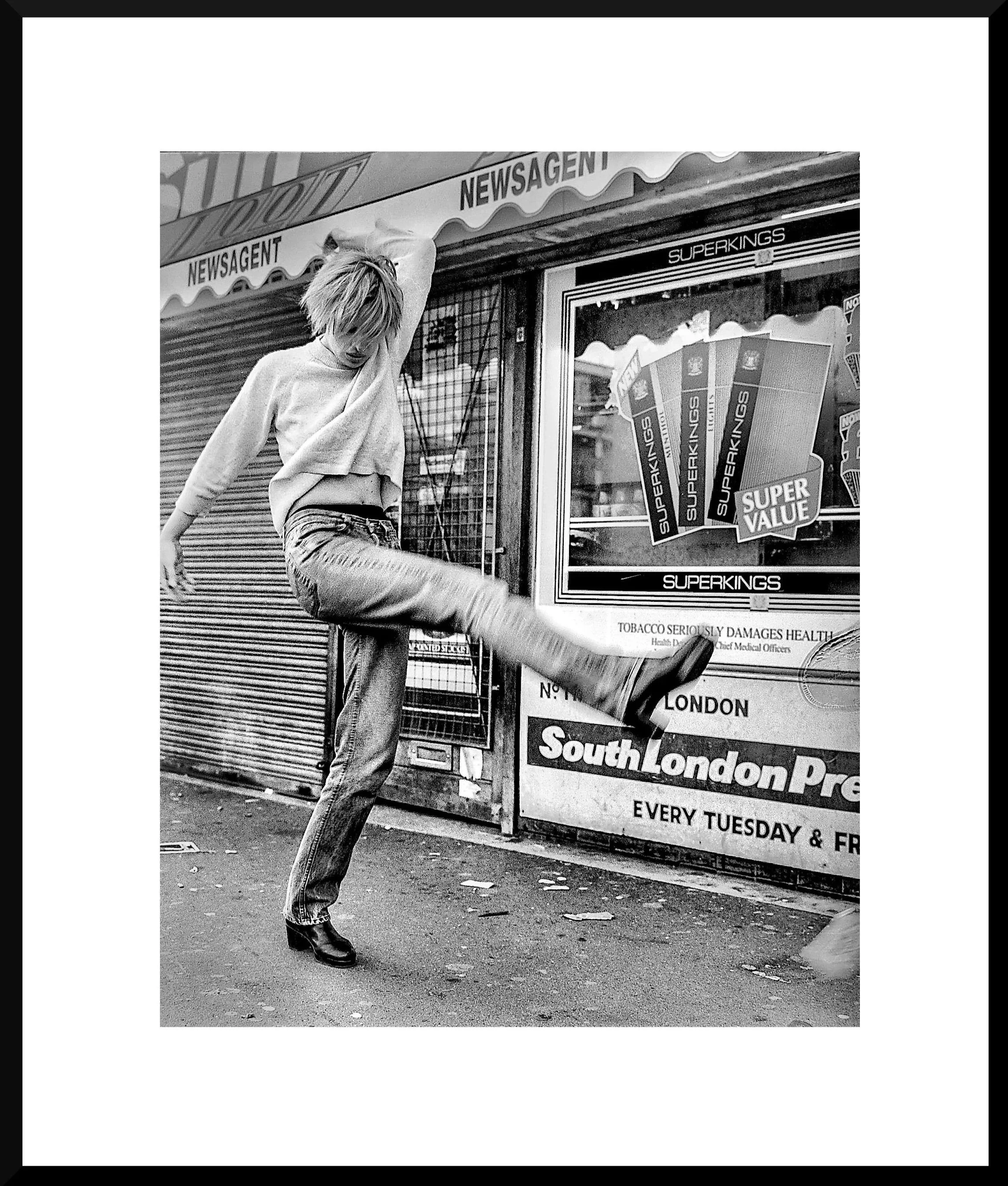 A woman in casual clothing, with her arm raised and face turned downward, is kicking a street-side shop shutter open in a street scene.