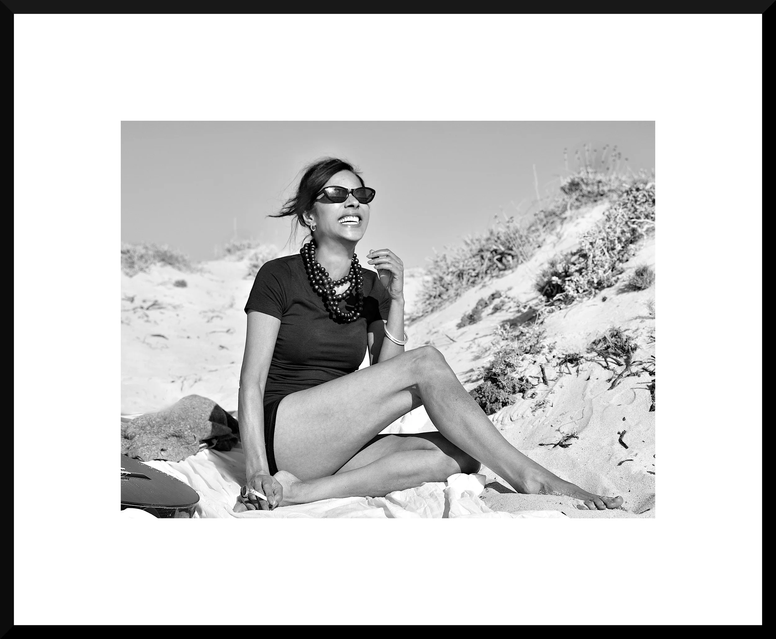 Woman in sunglasses and jewelry sitting on the sand at the beach, smiling and looking upward, with dunes and vegetation in the background.