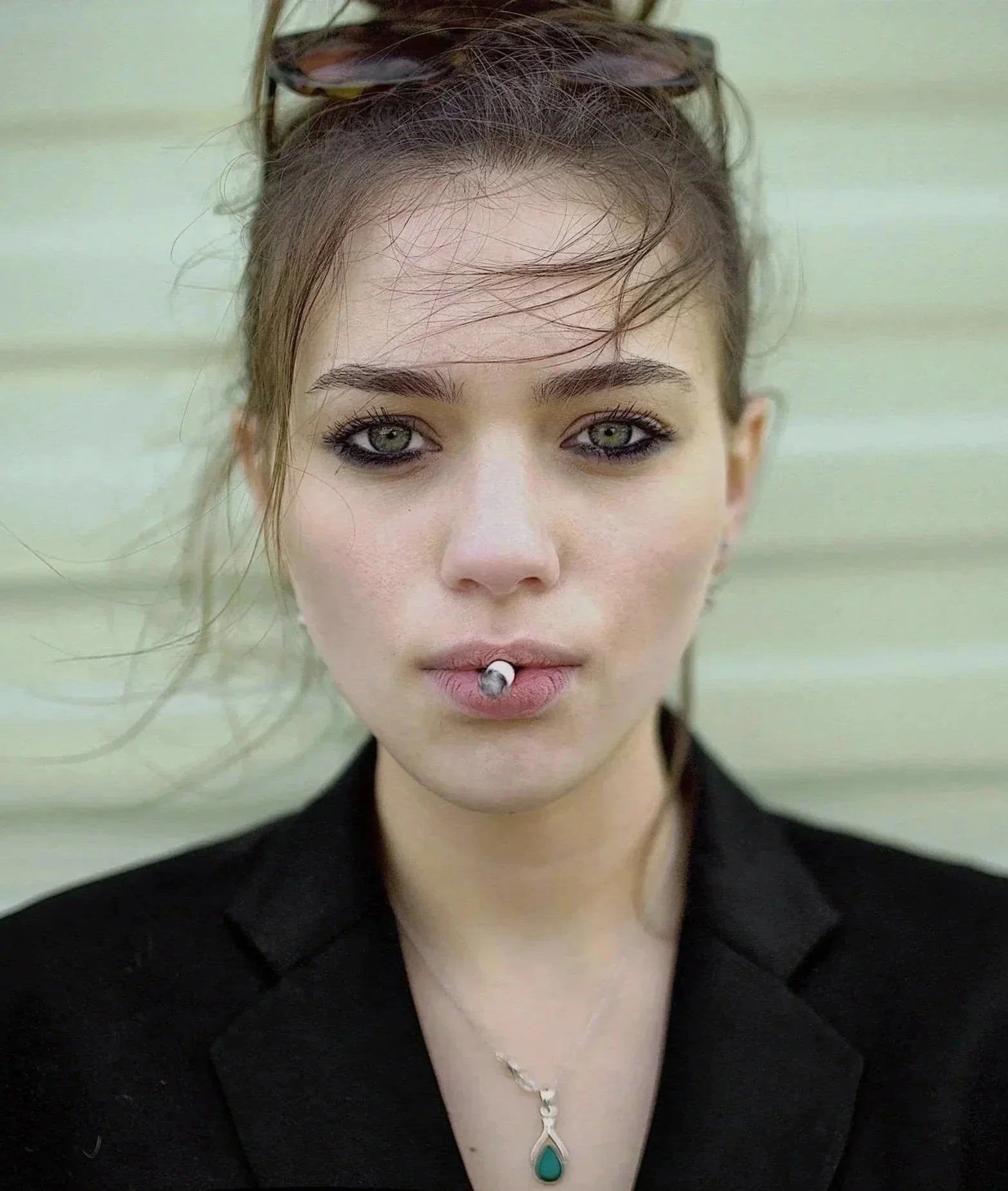 A young woman with fair skin, green eyes, and dark makeup, wearing glasses on her head and a black top. She has a vertical labret piercing with a silver ball and a teal pendant necklace. Her hair is styled loosely around her face, and she is standing against a pale, horizontally-striped background.
