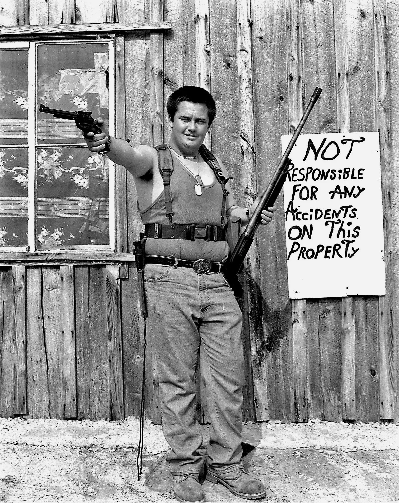 Black and white photograph of a young man standing outdoors in front of a wooden wall and window. He is holding a handgun pointed outward in his right hand and a large rifle in his left hand, with a serious expression. He is dressed in casual outdoor clothing, including a short-sleeved shirt, cargo pants, and boots. There is a sign on the wooden wall that says, "Not responsible for any accidents on this property."