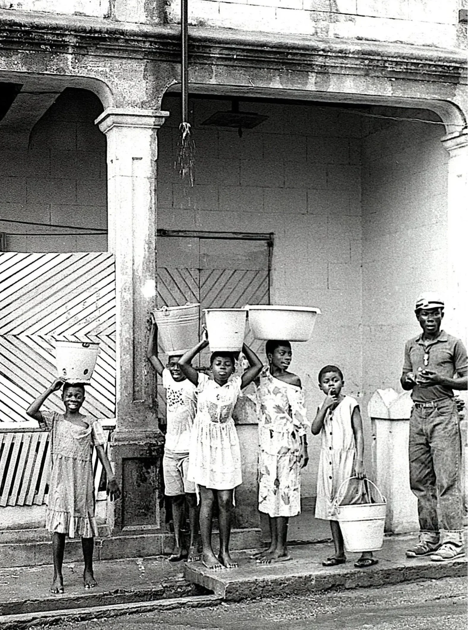 A group of children and an adult standing outside a building, some children carrying buckets on their heads, with a man standing nearby, all smiling or looking at the camera.