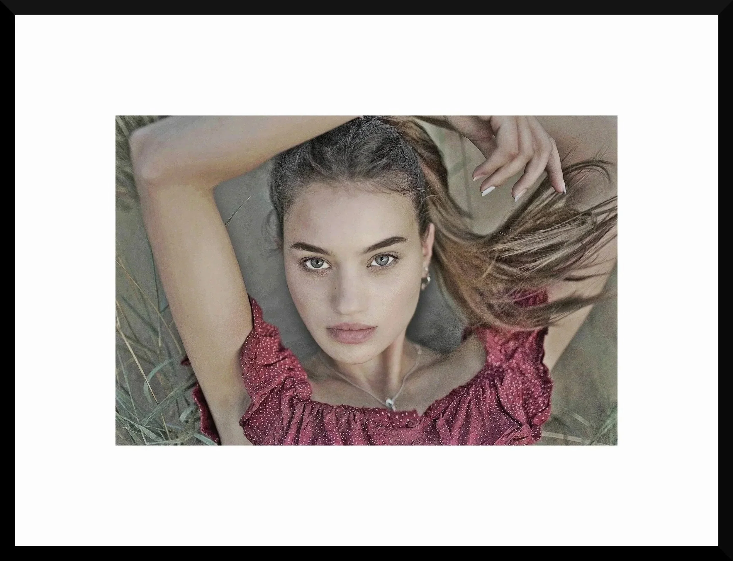 A young woman with light skin and blue eyes lying on the grass, wearing a red patterned blouse, looking up at the camera with her arms behind her head and long hair spread around her.