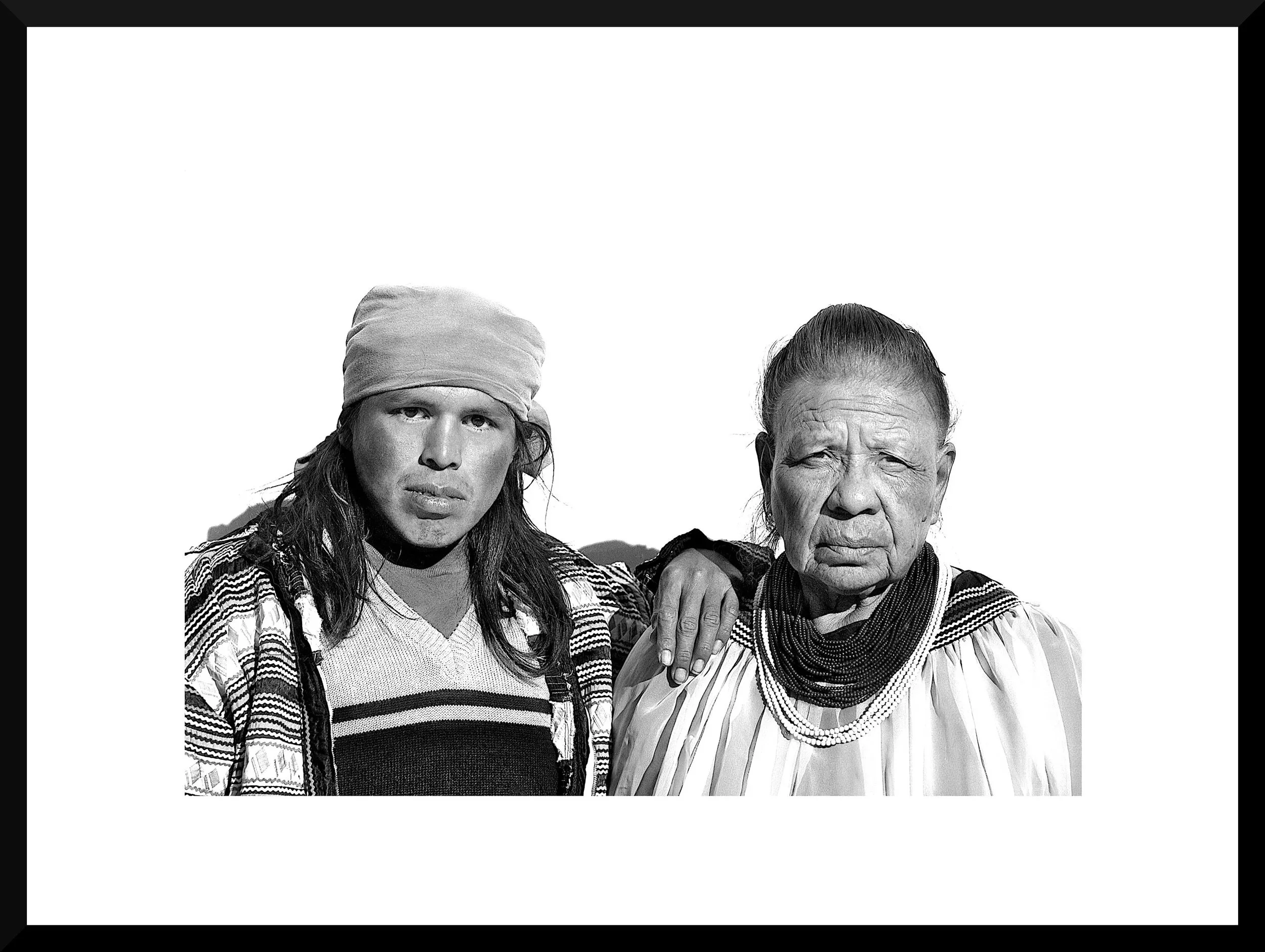 Black and white photo of a young woman with long hair wearing a headscarf and striped sweater, and an elderly woman with short hair wearing a patterned blouse and beaded necklaces, standing close together.