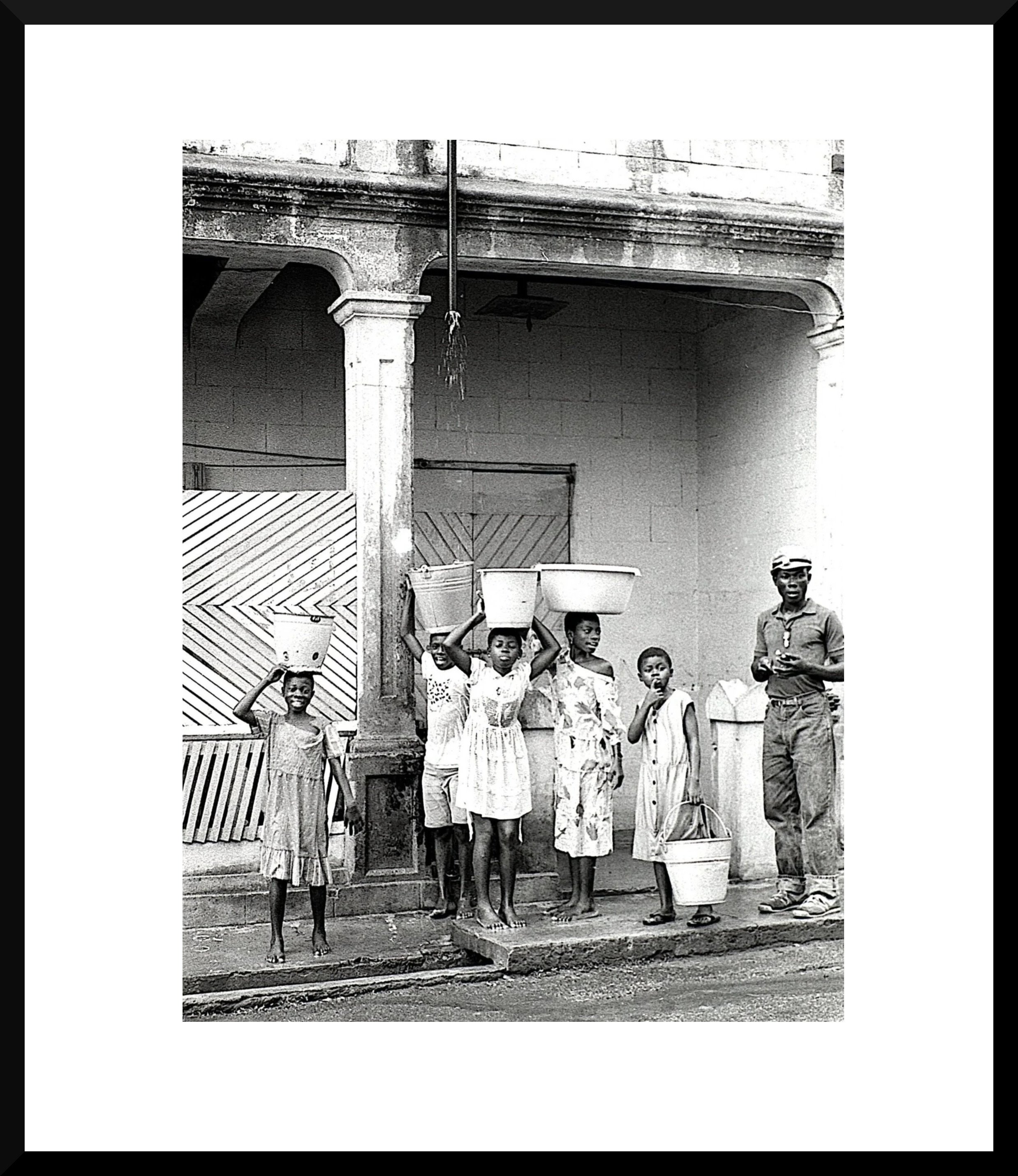Black and white photo of six children with buckets on their heads standing outside a building with columns, and an adult male standing to the right holding a cup.