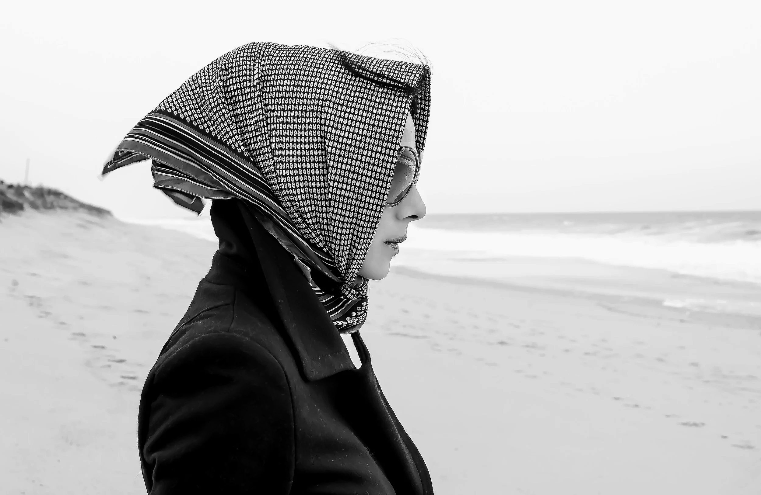 A woman wearing a patterned headscarf, sunglasses, and a dark jacket standing on a beach, with the ocean and sky in the background.