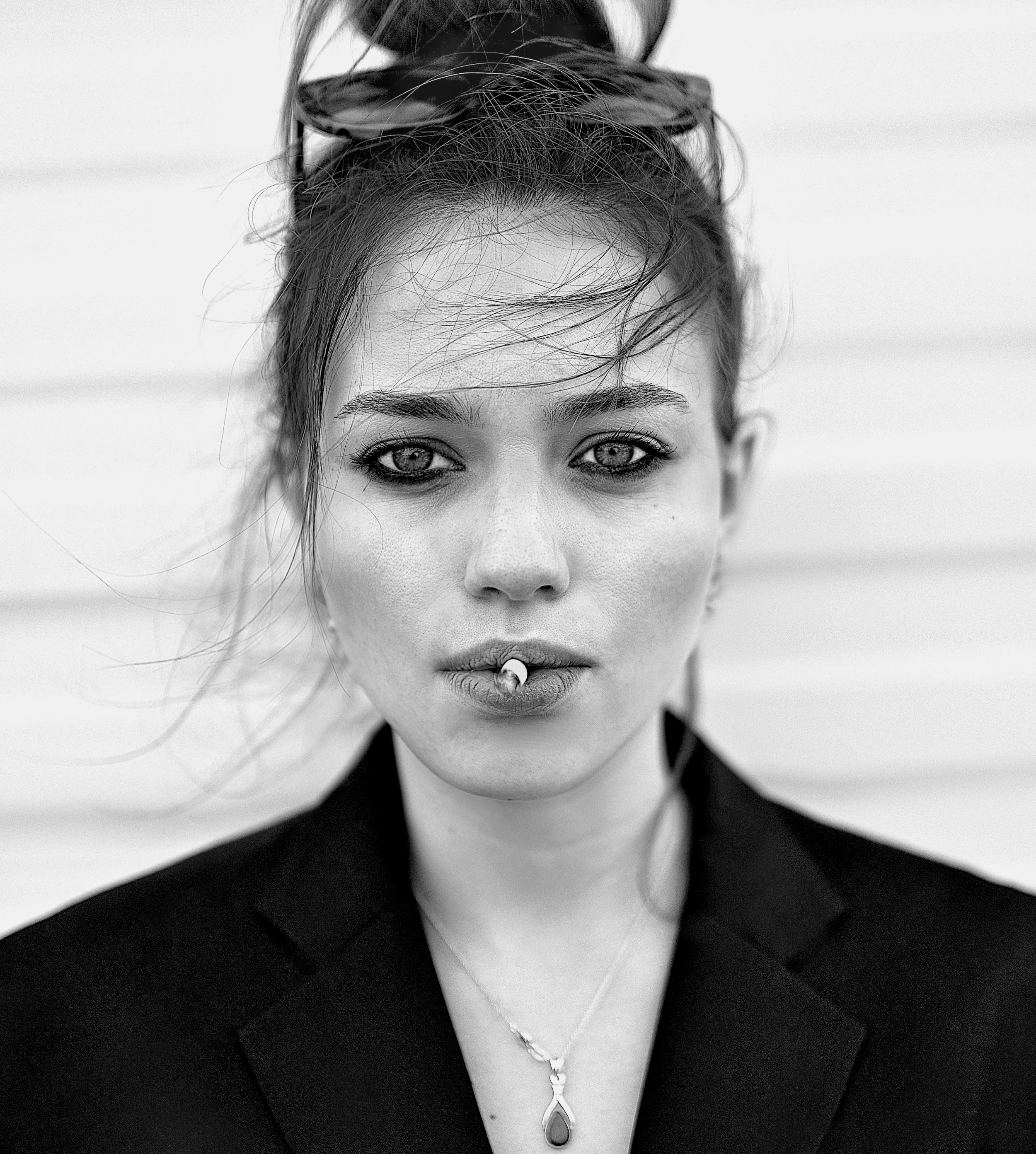 Black and white portrait of a woman with light-colored eyes, messy hair, and a cigarette in her mouth, wearing a dark blazer and a necklace.