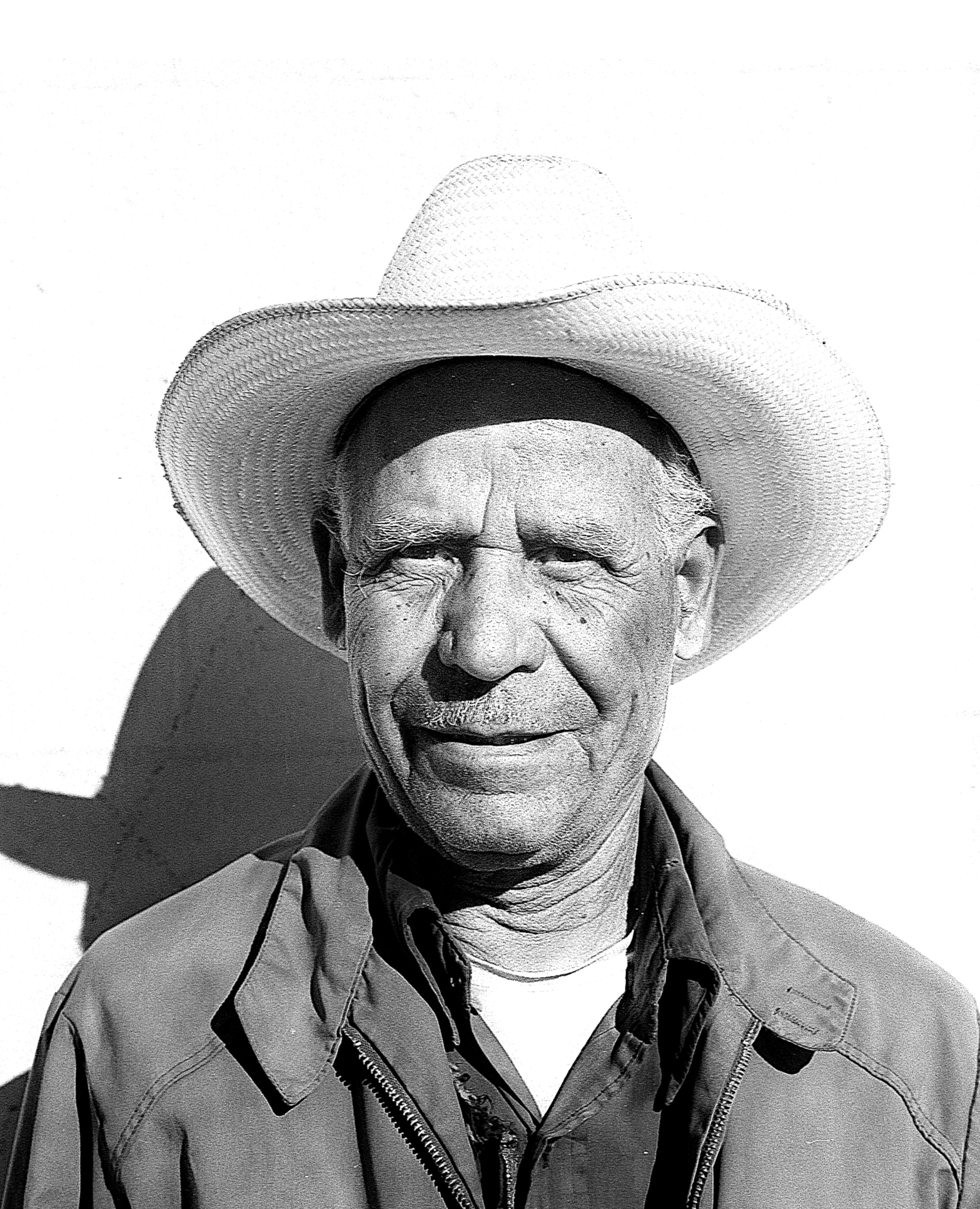An elderly man with a cowboy hat, white hair, and glasses. He is dressed in a jacket and shirts, standing outdoors with a shadow cast on his face.