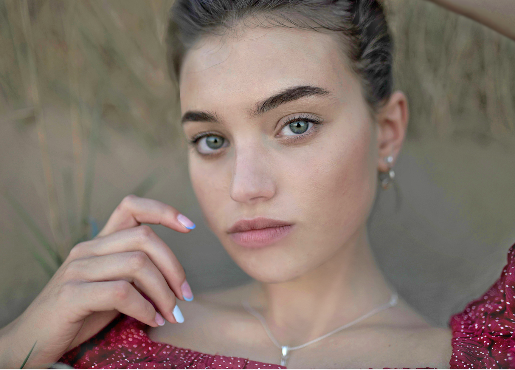 Close-up of a young woman with blue eyes, brown hair, and fair skin lying outdoors among tall grass, wearing a red dress with white polka dots, silver earrings, and a necklace, with her hand near her face.