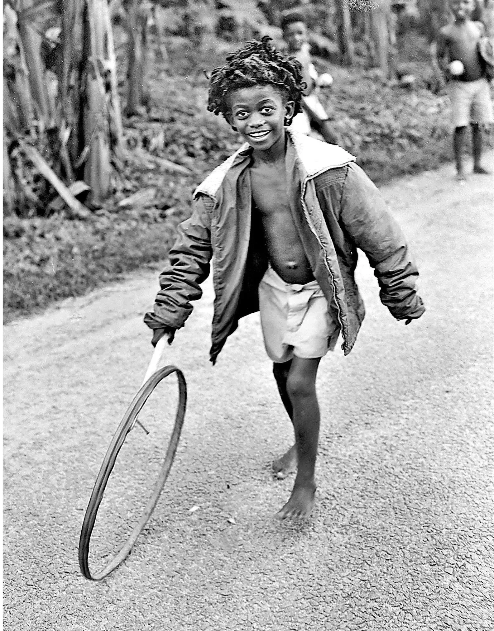 A young girl is walking barefoot down a dirt or gravel pathway in a rural setting, smiling at the camera with her arms outstretched. She is wearing a partially unzipped jacket and shorts. Other children are visible in the background among trees and foliage.