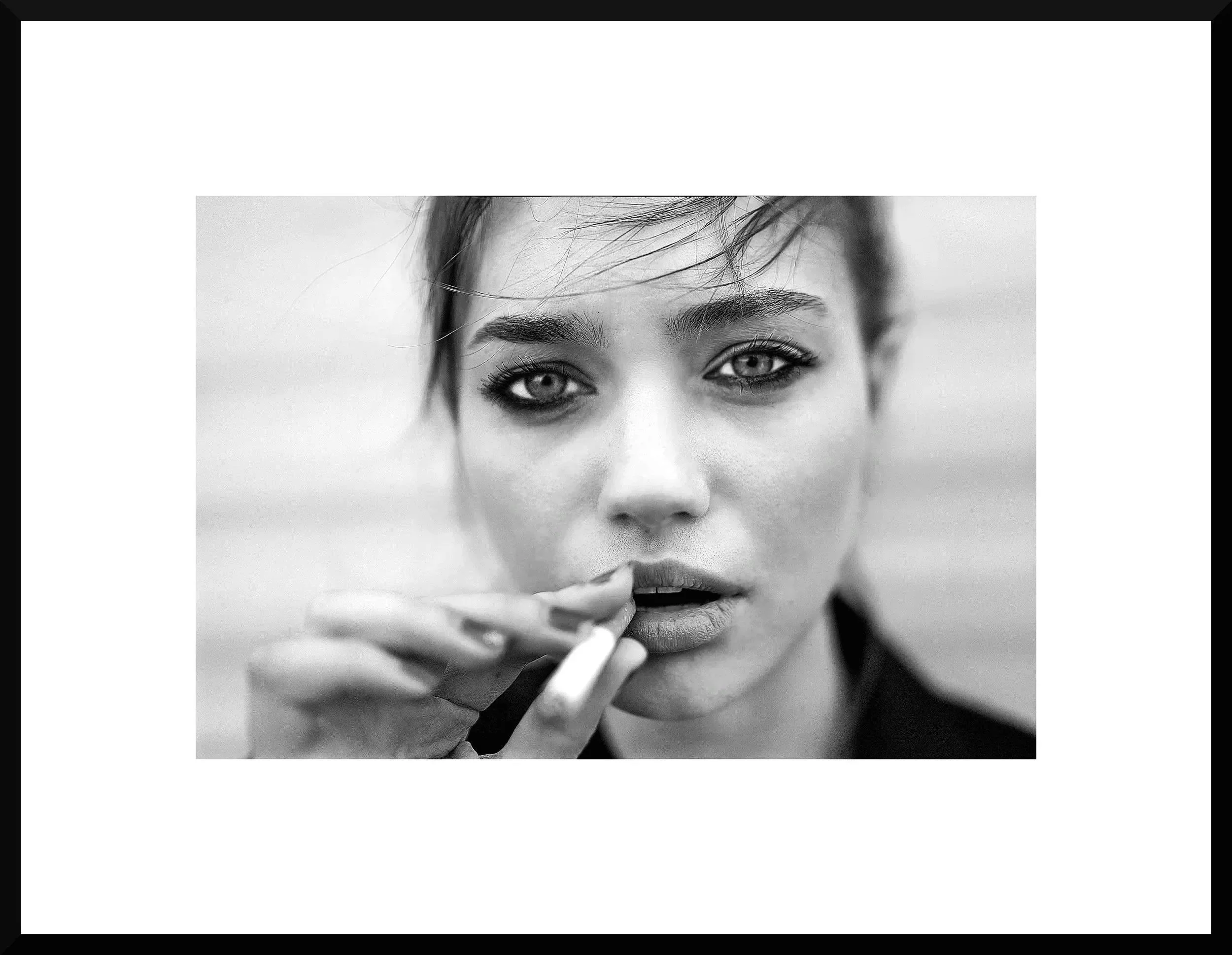 Close-up black and white photo of a woman with light-colored eyes holding a cigarette near her lips. Her gaze is directed toward the camera, and her hair partially covers her forehead.
