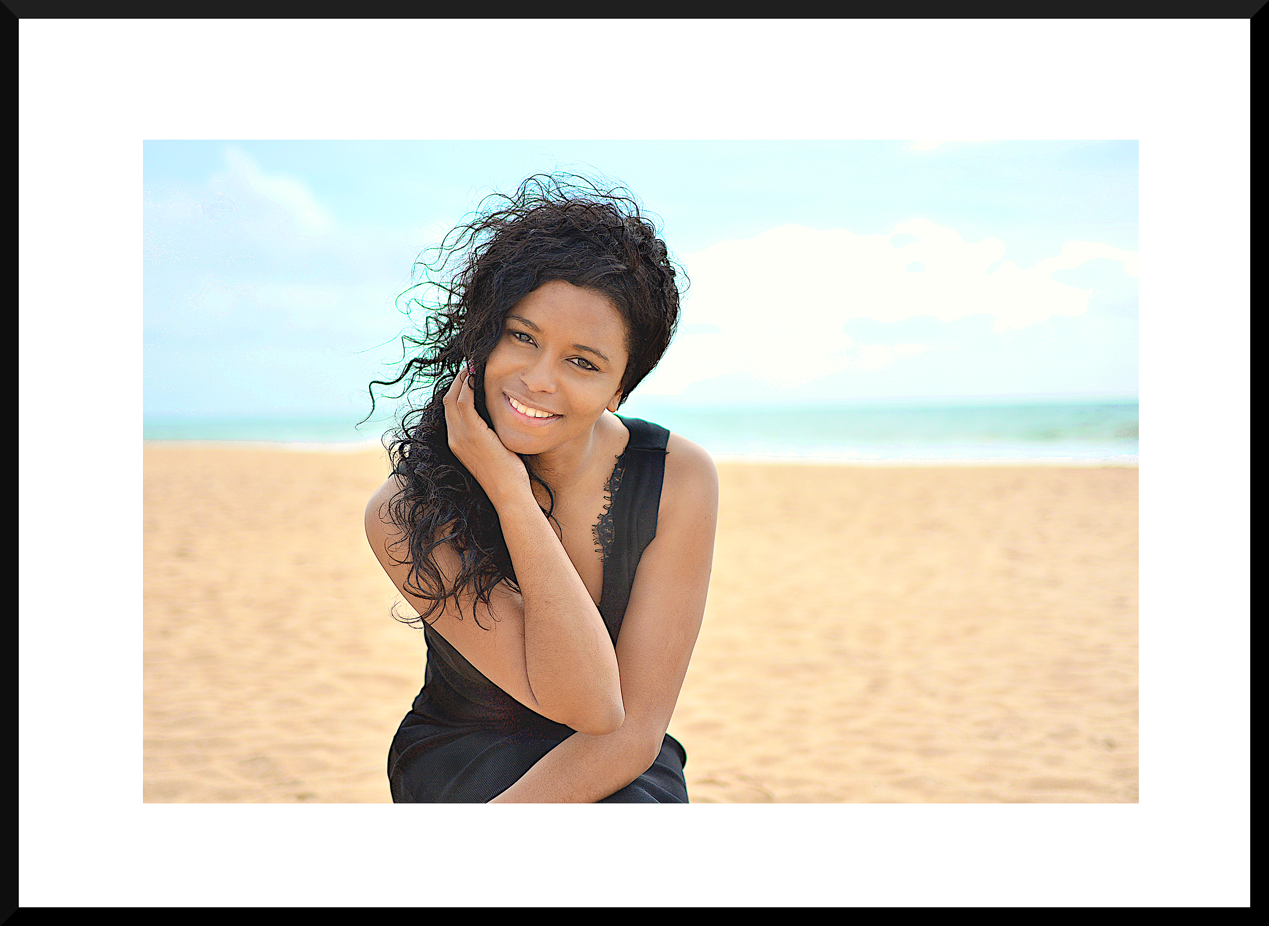 A woman with dark curly hair, smiling, posing outdoors on a beach with sand and water in the background.