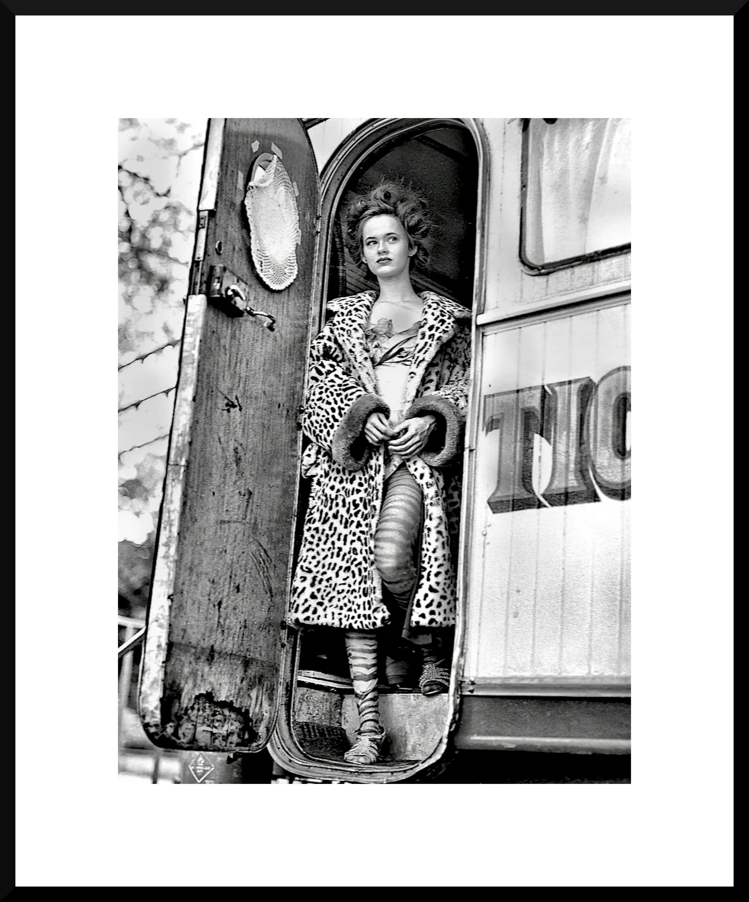 A woman in vintage clothing standing in the doorway of a caravan or trailer, wearing a leopard print coat and striped tights, with her hair styled in a wavy, voluminous fashion.