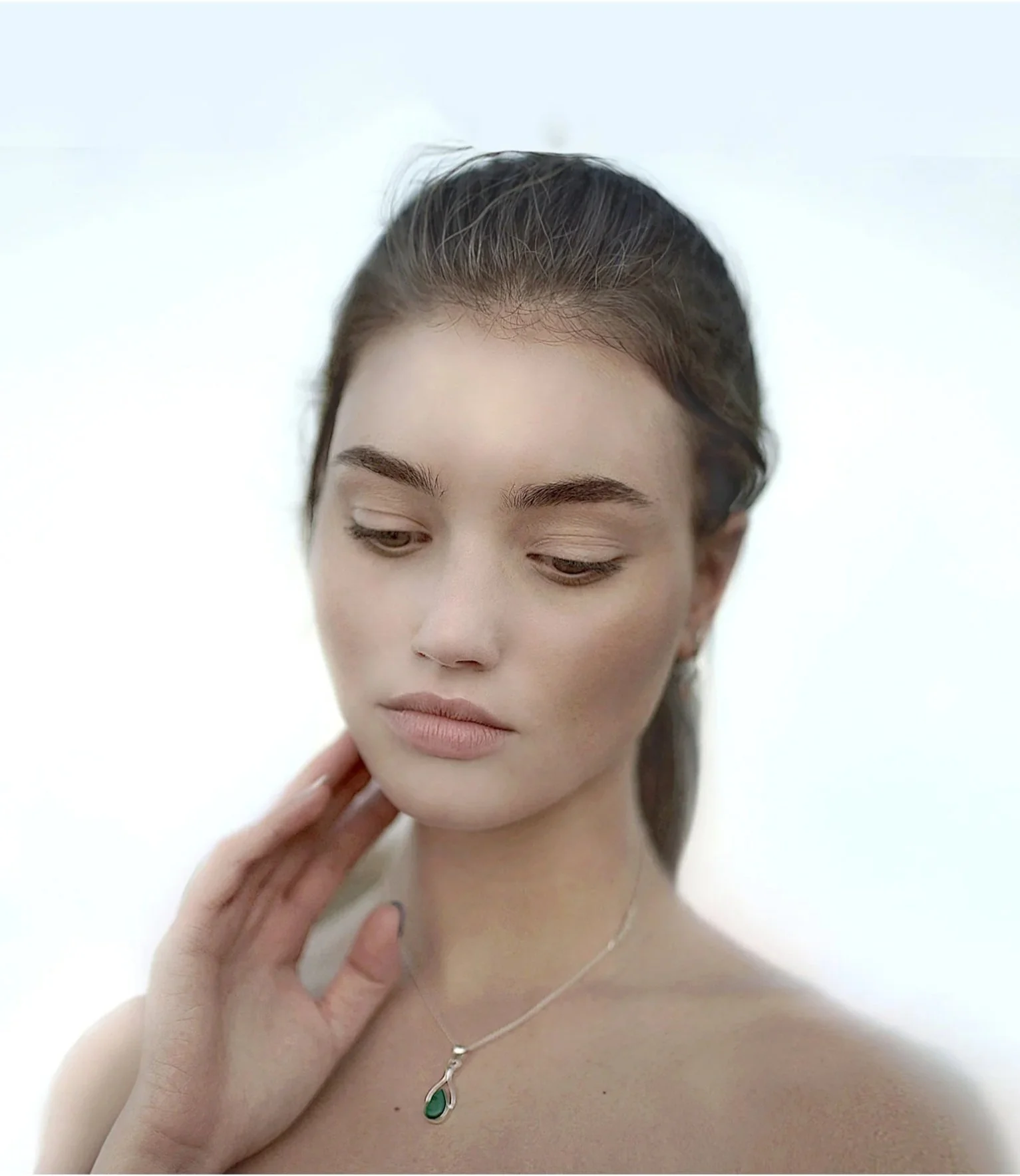 A woman with brown hair, wearing a silver necklace with a green pendant, looks down with her eyes closed, touching her neck with her left hand.