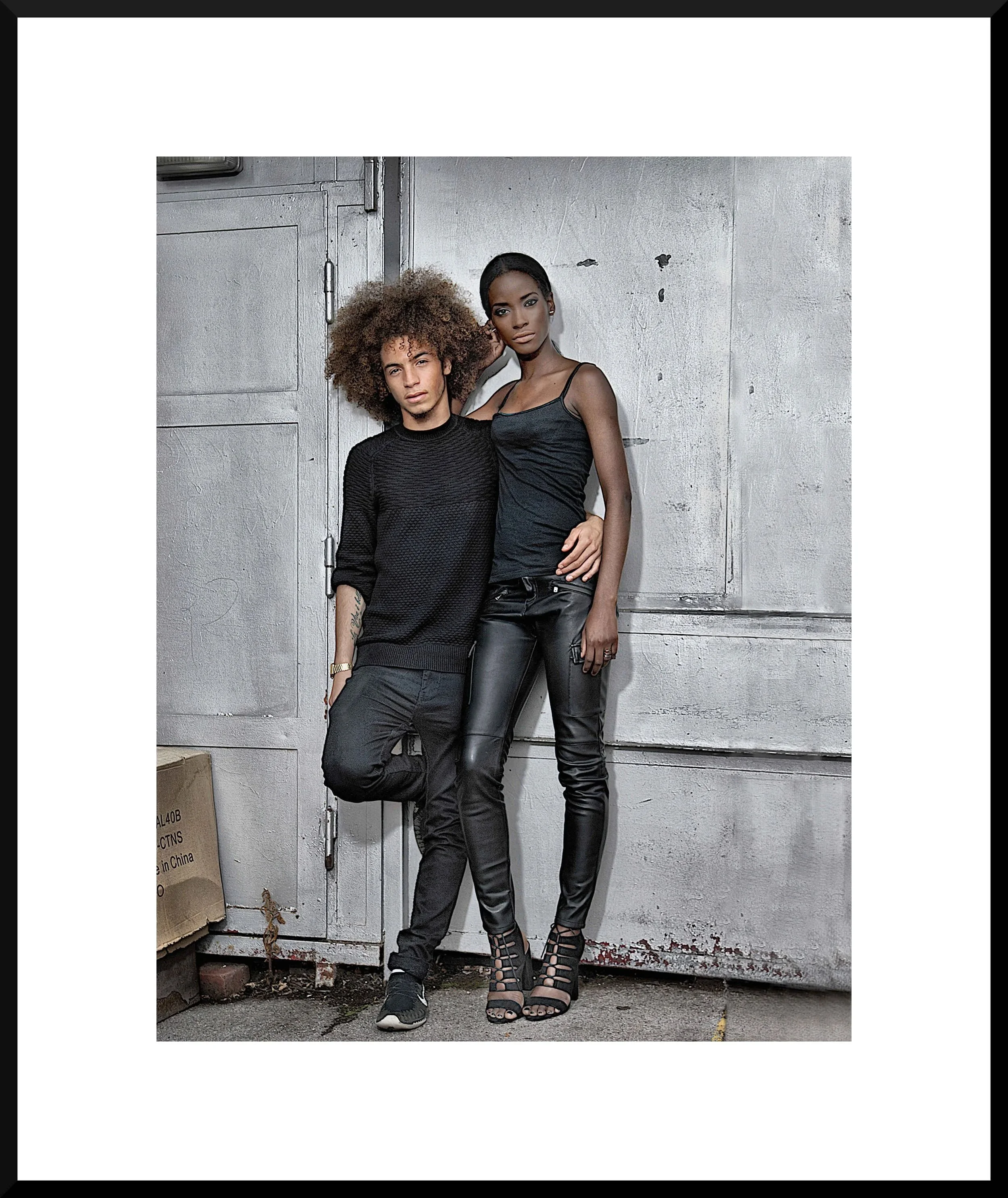 A young man and woman dressed in black clothing standing closely against a textured gray wall.