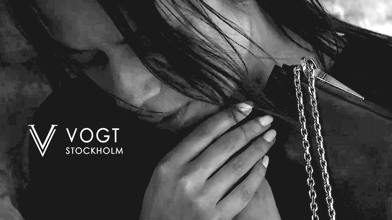 Black and white photo of a woman with long dark hair, holding a lipstick near her lips, with chains hanging from her clothing, and a logo that reads 'V VOGT STOCKHOLM' on the lower left corner.