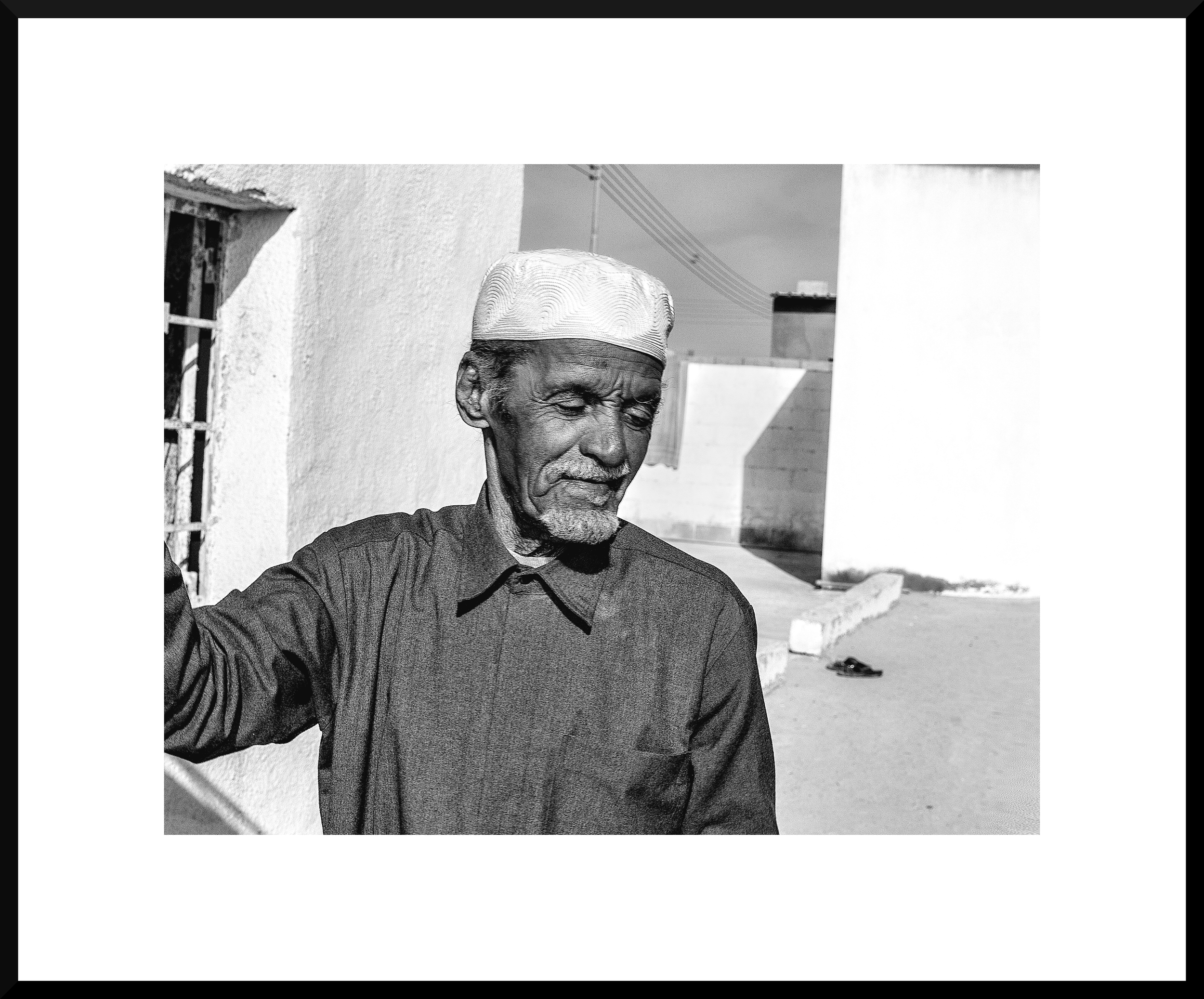 A black and white photo of an elderly man wearing a traditional cap and a jacket, standing outdoors near a white wall, with a clear sky and some electrical wires in the background.