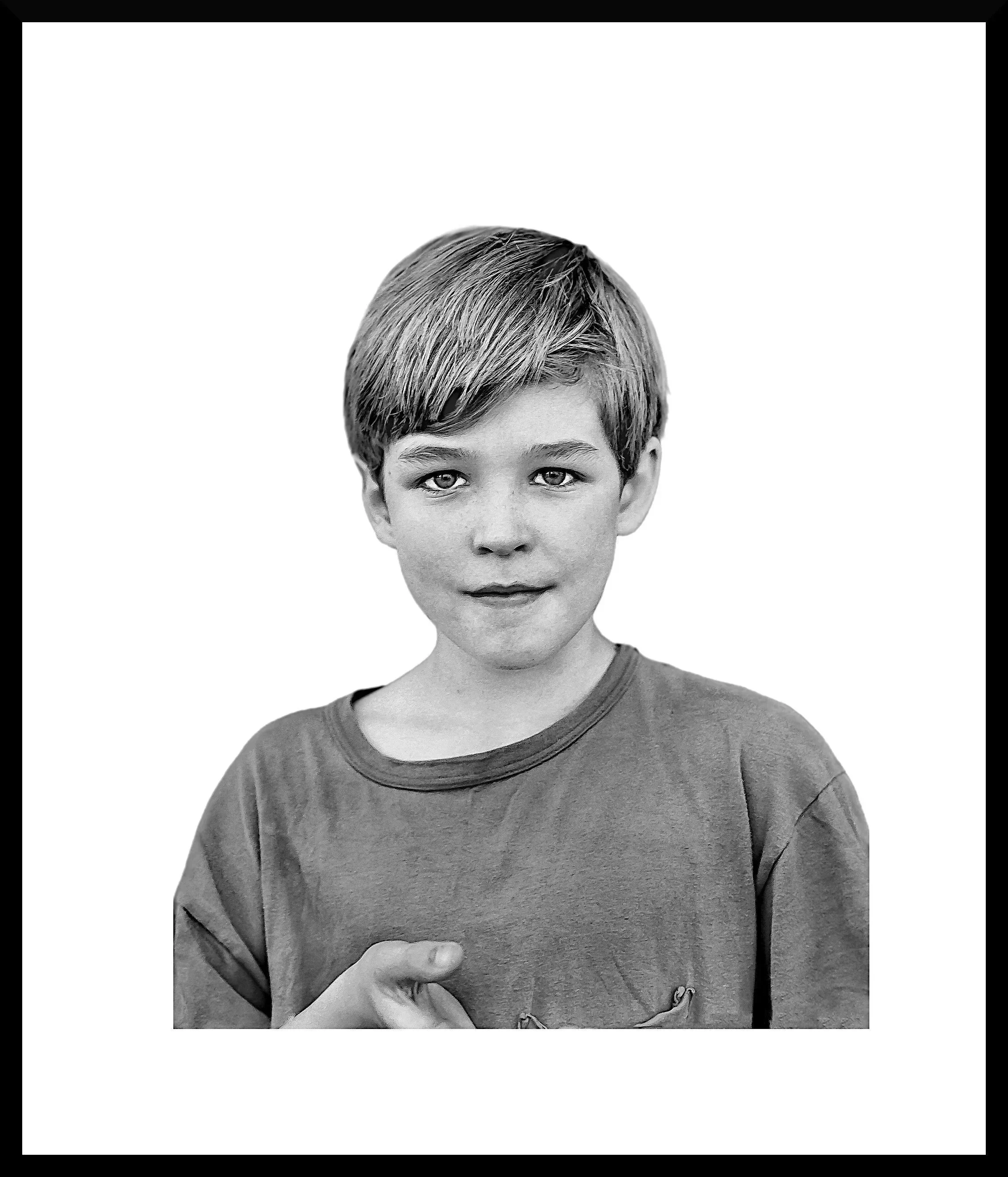 Black and white photo of a young boy with short hair, wearing a T-shirt, looking directly at the camera with a neutral expression, and his hand raised near his chest.