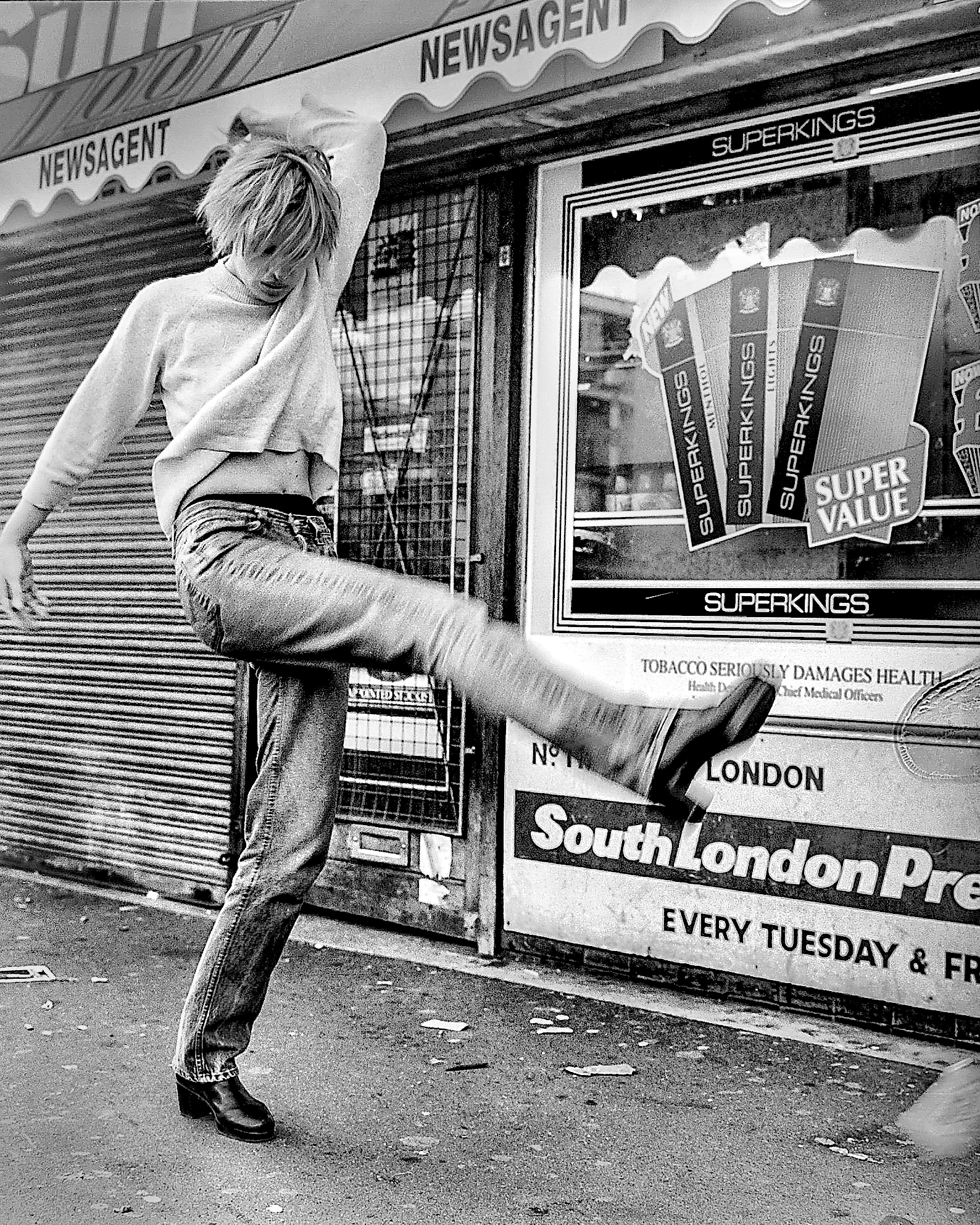 A person kicking a newspaper stand outside a shop with a sign that reads "NEWSAGENT". The person has short hair and is wearing a loose sweatshirt, jeans, and boots. The image is black and white.