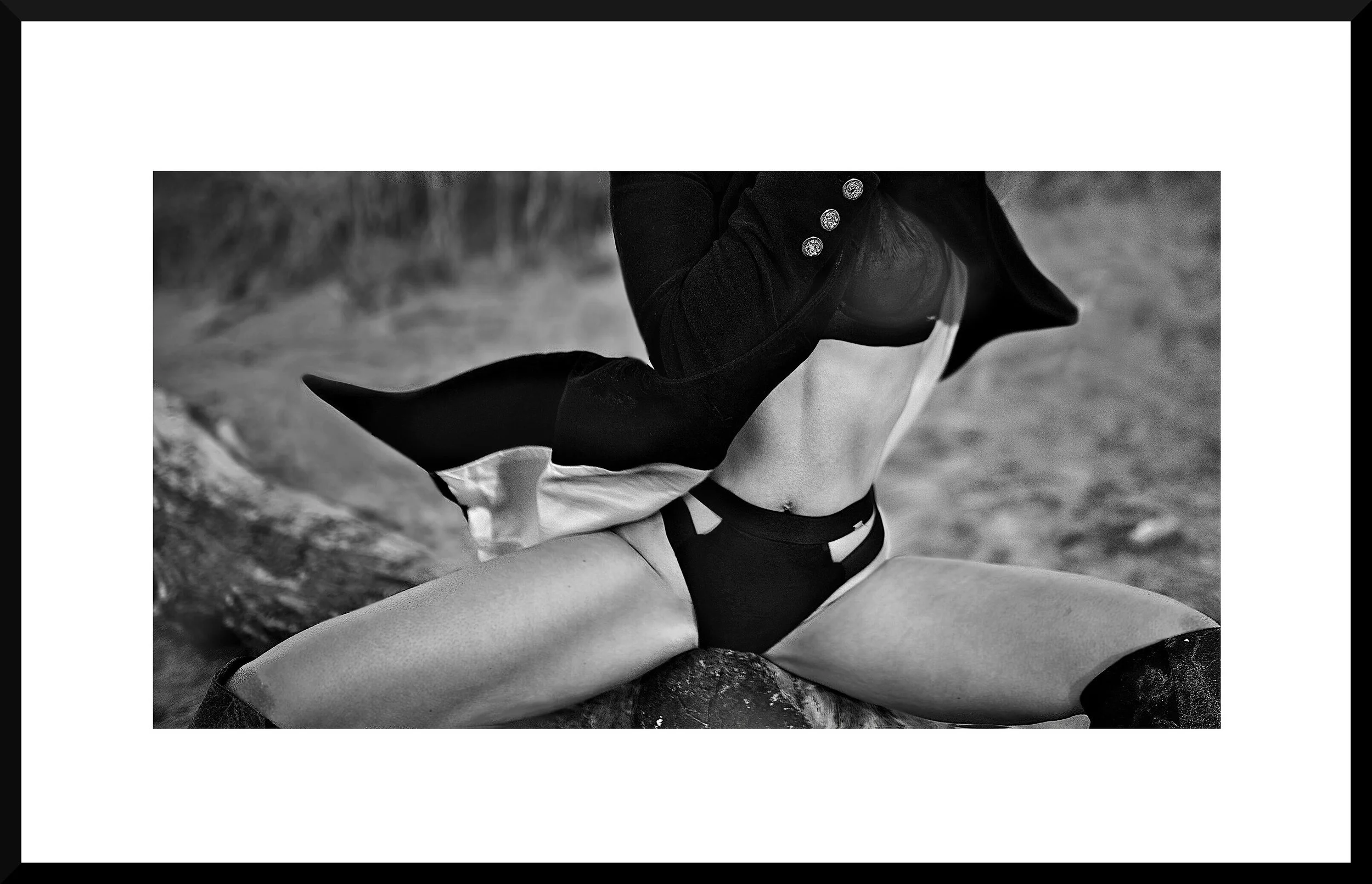 A person in black and white lingerie and thigh-high boots poses outdoors on a large rock, with their jacket pulled open to reveal their torso and part of their legs.