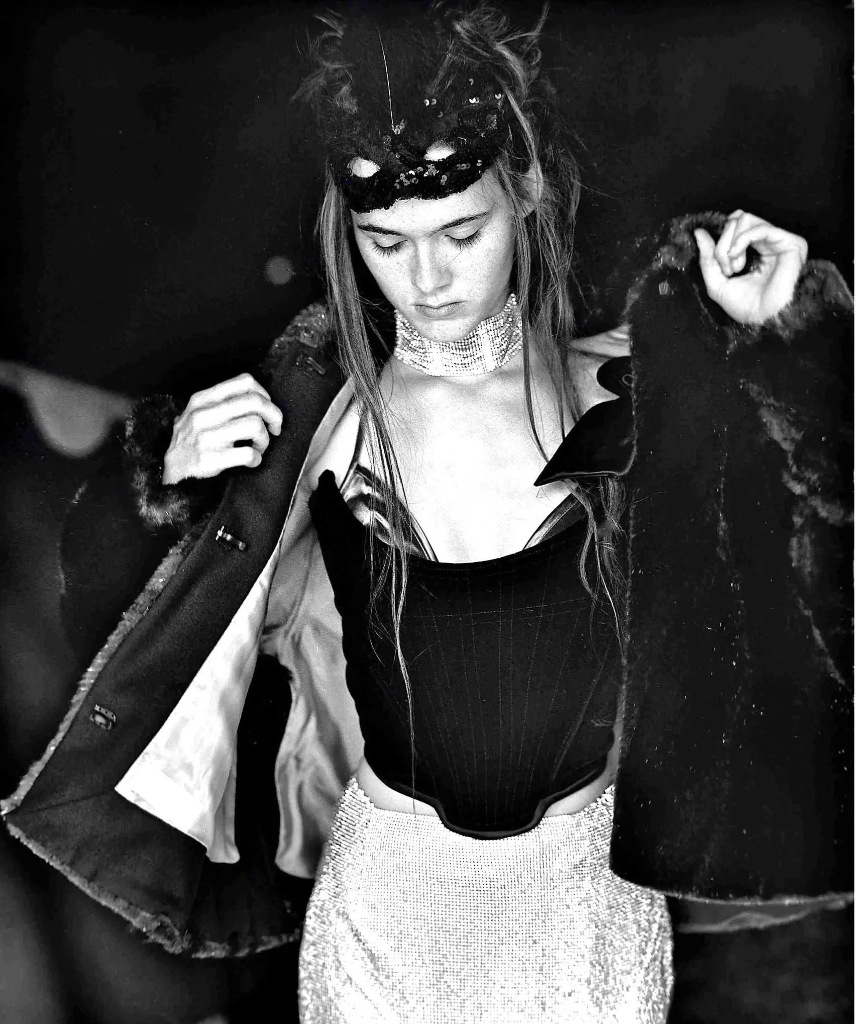 A black-and-white photograph of a young woman with long hair, wearing a sequined top, a braided jacket with fur cuffs, a choker necklace, and a pair of sunglasses lowered to reveal her face, against a dark background.