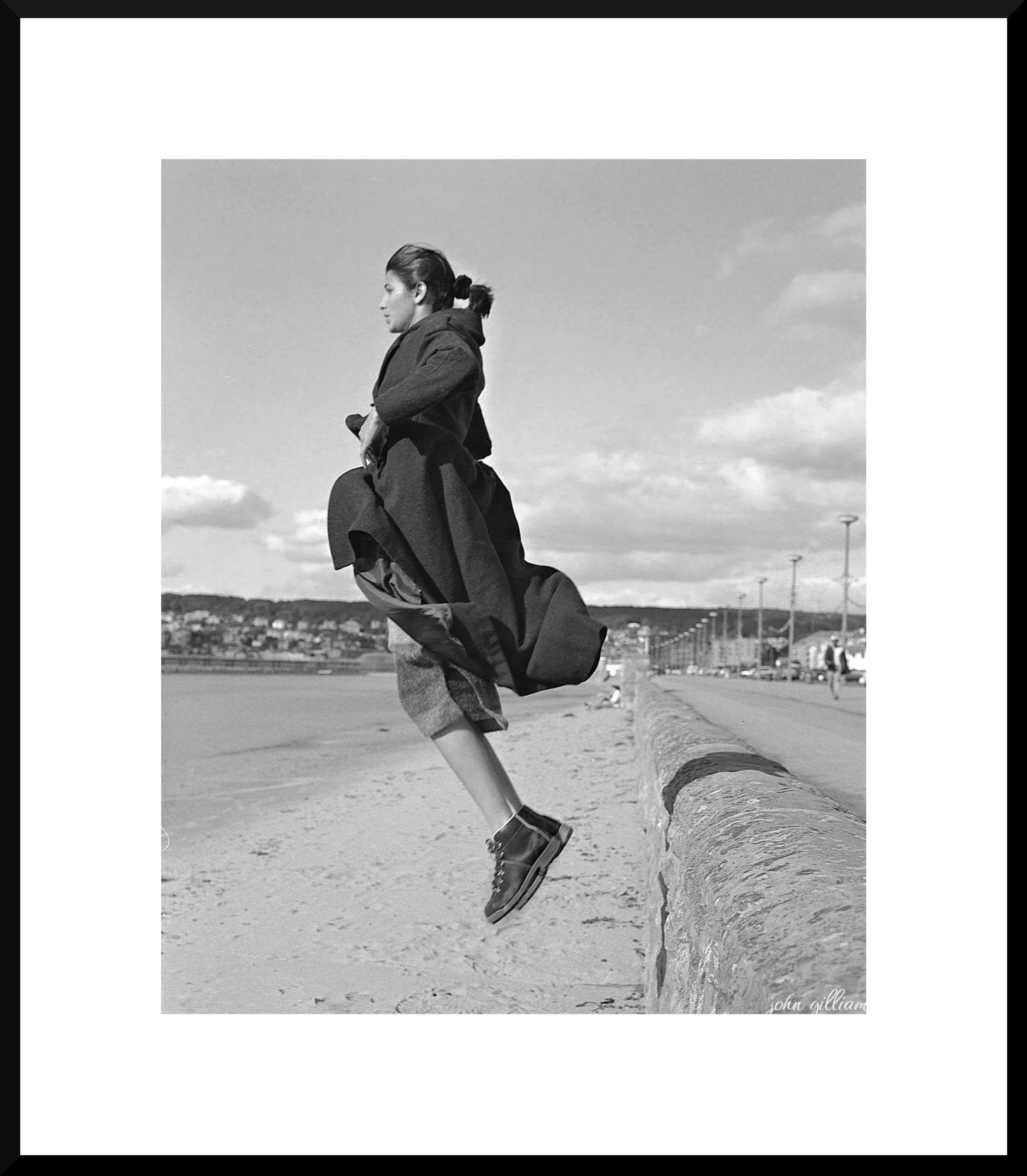 A black-and-white photo of a woman jumping over a brick barrier on a beach, with the ocean and a cloudy sky in the background.