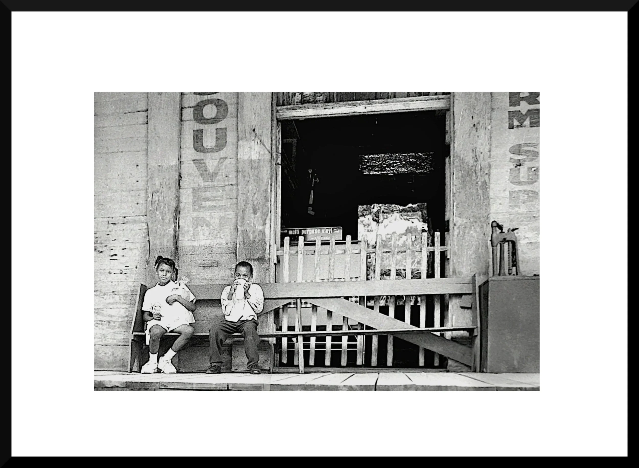Two children sitting on a bench in front of a rustic wooden building with an open doorway and a small fenced area.