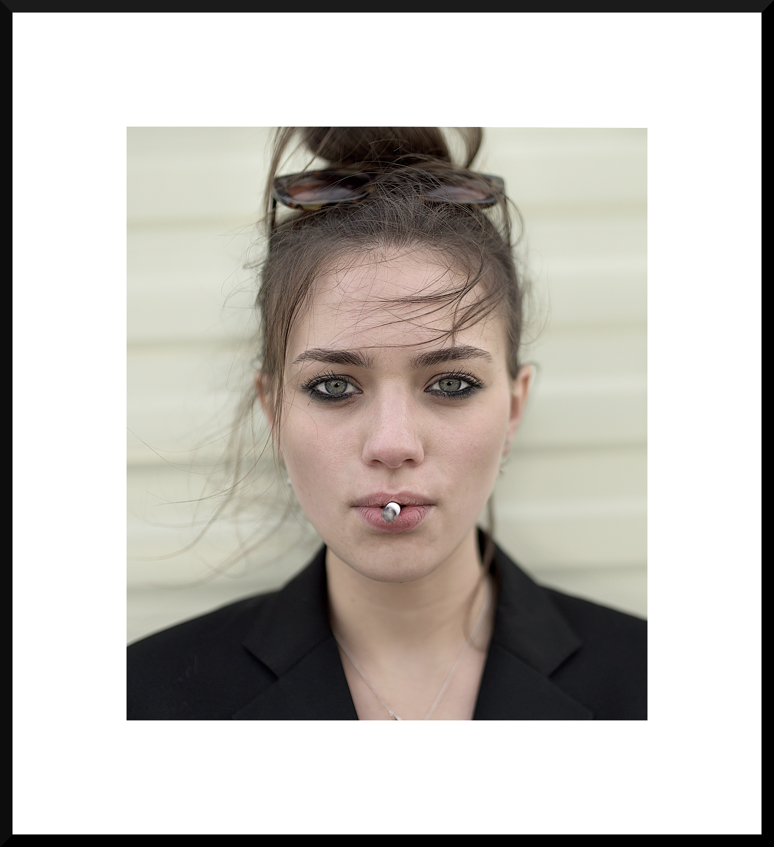 Portrait of a woman with light hair in a bun, wearing sunglasses on her head, smoking a cigarette, and wearing a black blazer, against a light-colored background.